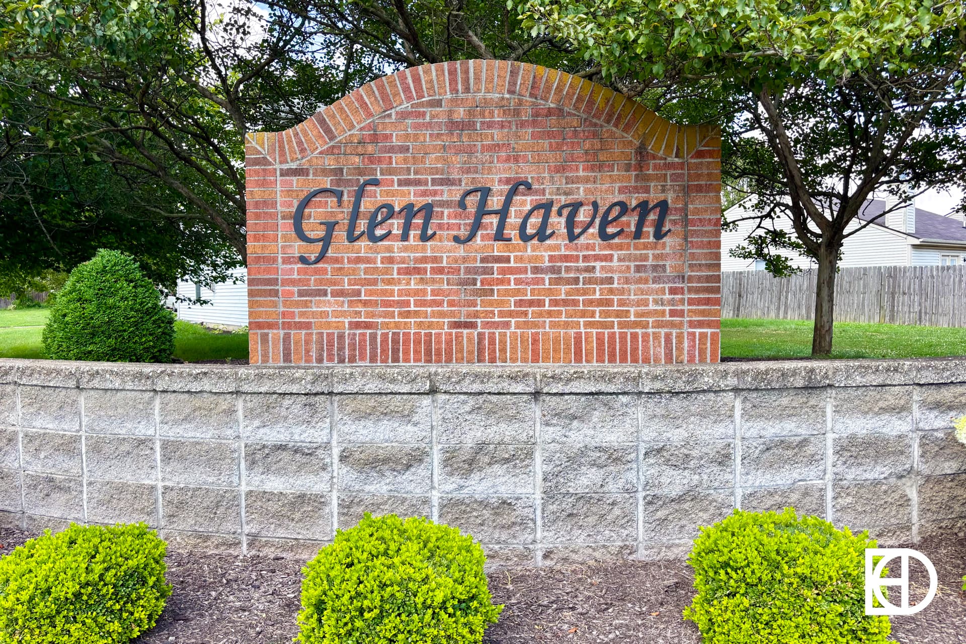 A brick and stone entrance sign reads Glen Haven in cursive text, surrounded by green bushes, grass, and trees, with houses and a wooden fence in the background.