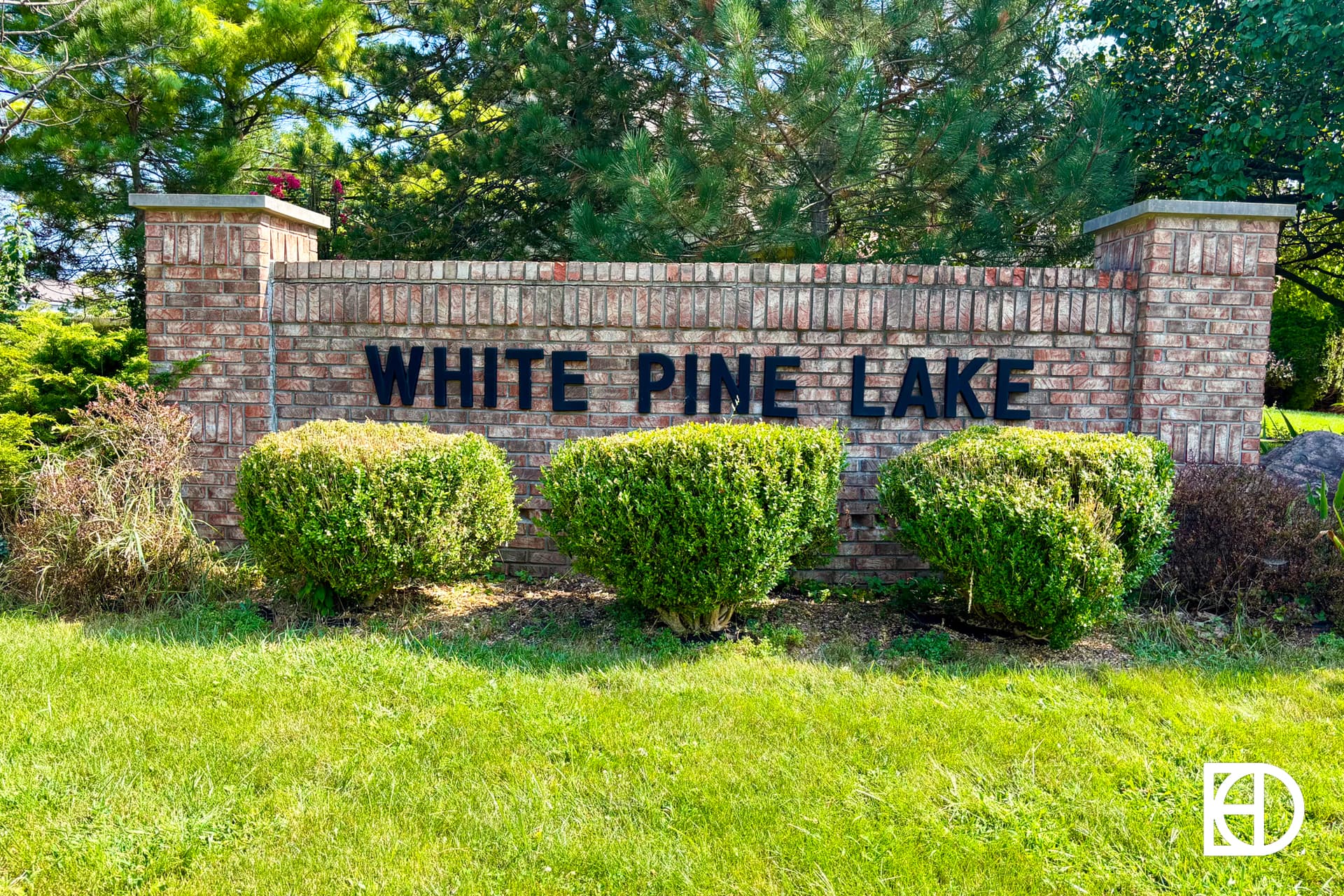 A brick sign with black letters reads WHITE PINE LAKE, surrounded by green bushes and trees, with grass in the foreground and a white KD logo in the lower right corner.