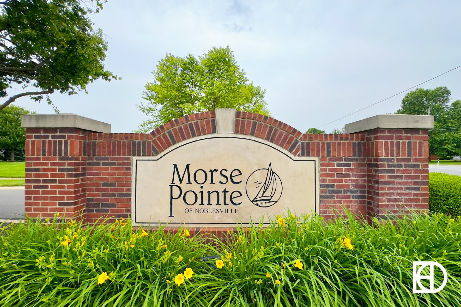 A brick entrance sign reads Morse Pointe of Noblesville with a sailboat graphic, surrounded by yellow flowers and green landscaping.