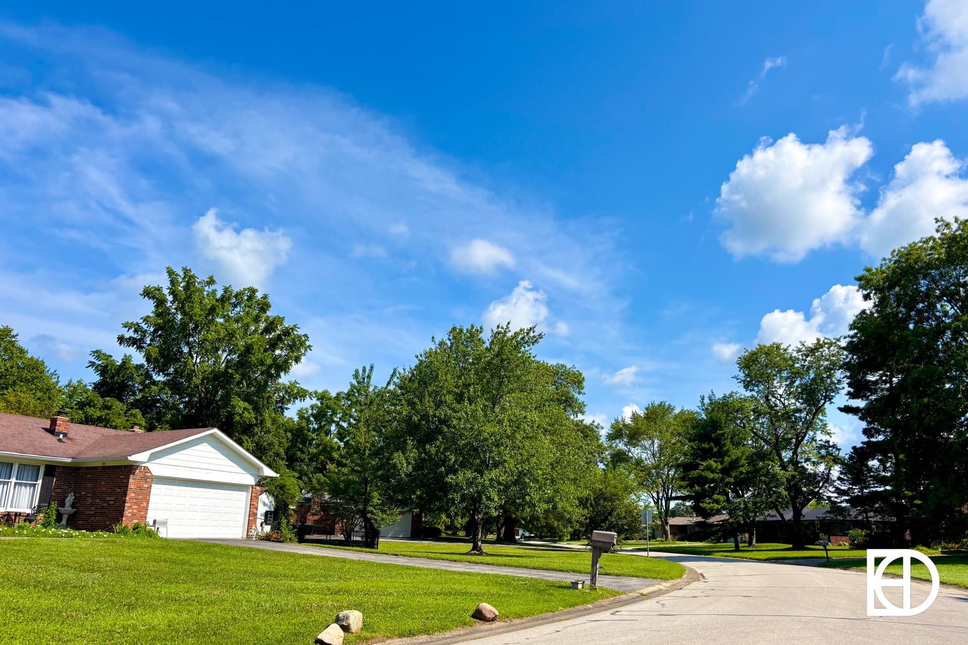 A quiet suburban street with single-story brick houses, green lawns, large leafy trees, and a bright blue sky with scattered clouds. A mailbox stands by the roadside under the sunlight.