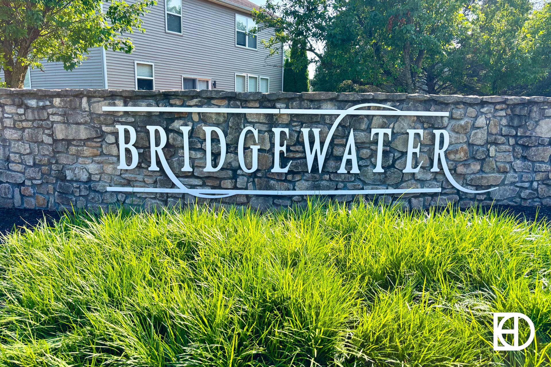 A stone wall with the word BRIDGEWATER written in large white letters, surrounded by green grass and trees, with a house partially visible in the background.