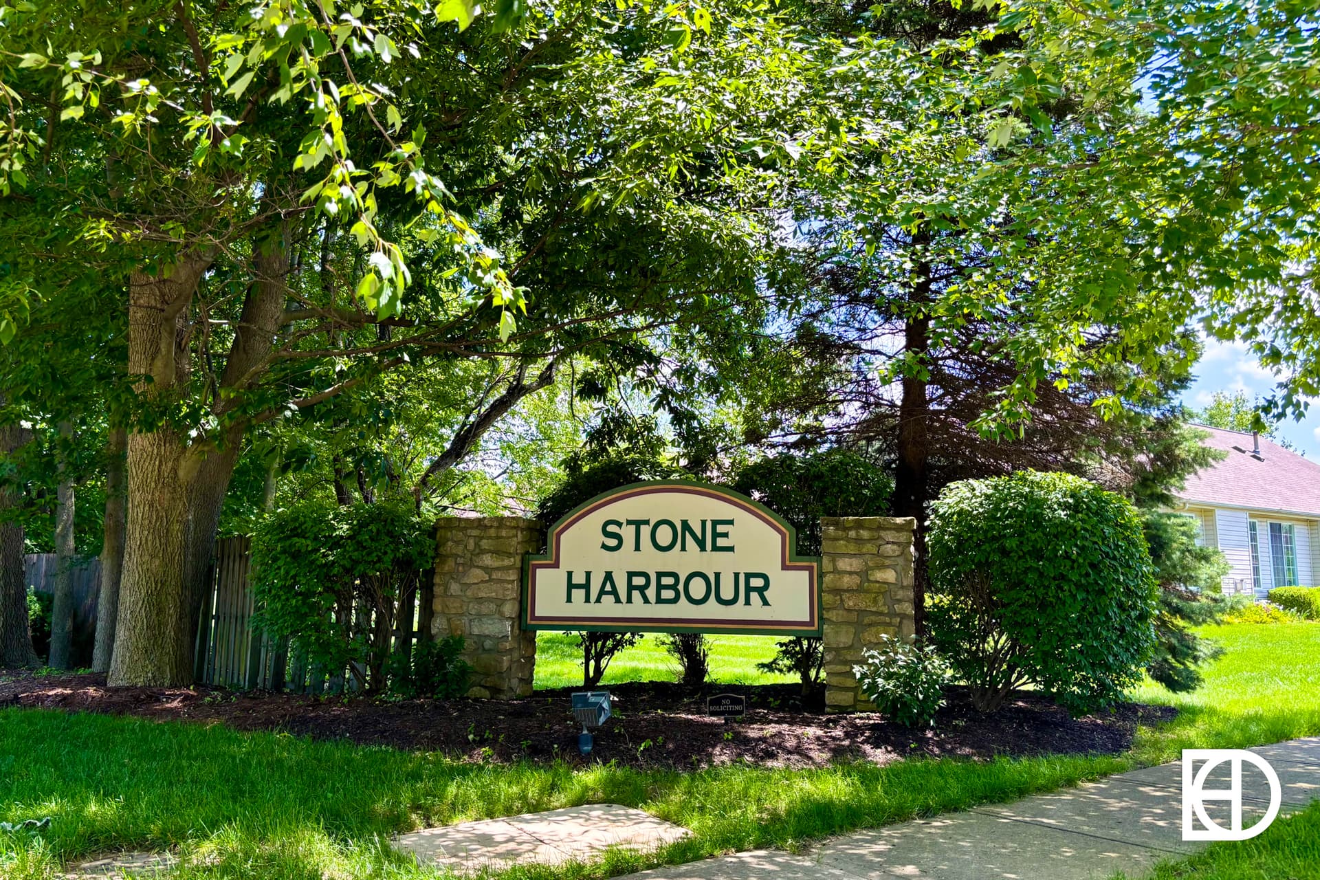 A stone and wood sign reading “Stone Harbour” stands among green bushes and trees beside a sidewalk, with part of a house visible in the background on a sunny day.