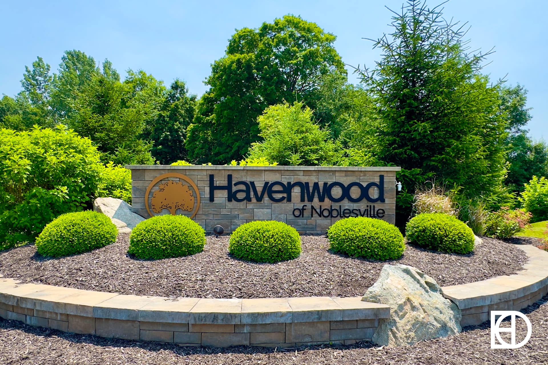 A welcoming entrance sign for Havenwood of Noblesville, featuring a wooden structure with the name, surrounded by trees, shrubs, and a landscaped pathway with mulch and stones. The sky is clear and sunny, with a white KD logo in the bottom right corner.
