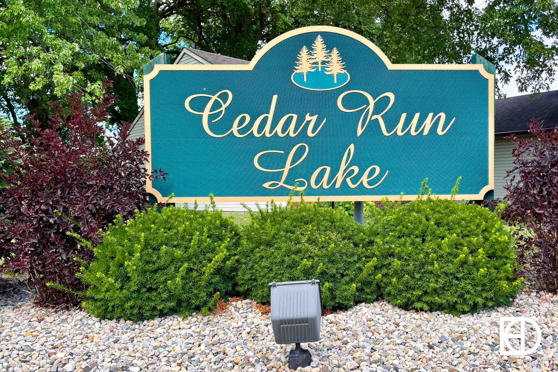A green and gold sign reads Cedar Run Lake with tree graphics above the text. The sign is surrounded by bushes and rocks, with trees and a building in the background.