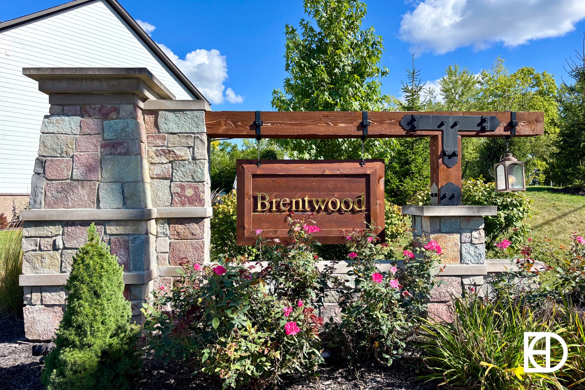 A decorative stone and wood sign reading Brentwood stands among blooming pink roses and greenery, with a lantern hanging on the right. Blue sky and a house roof are visible in the background.