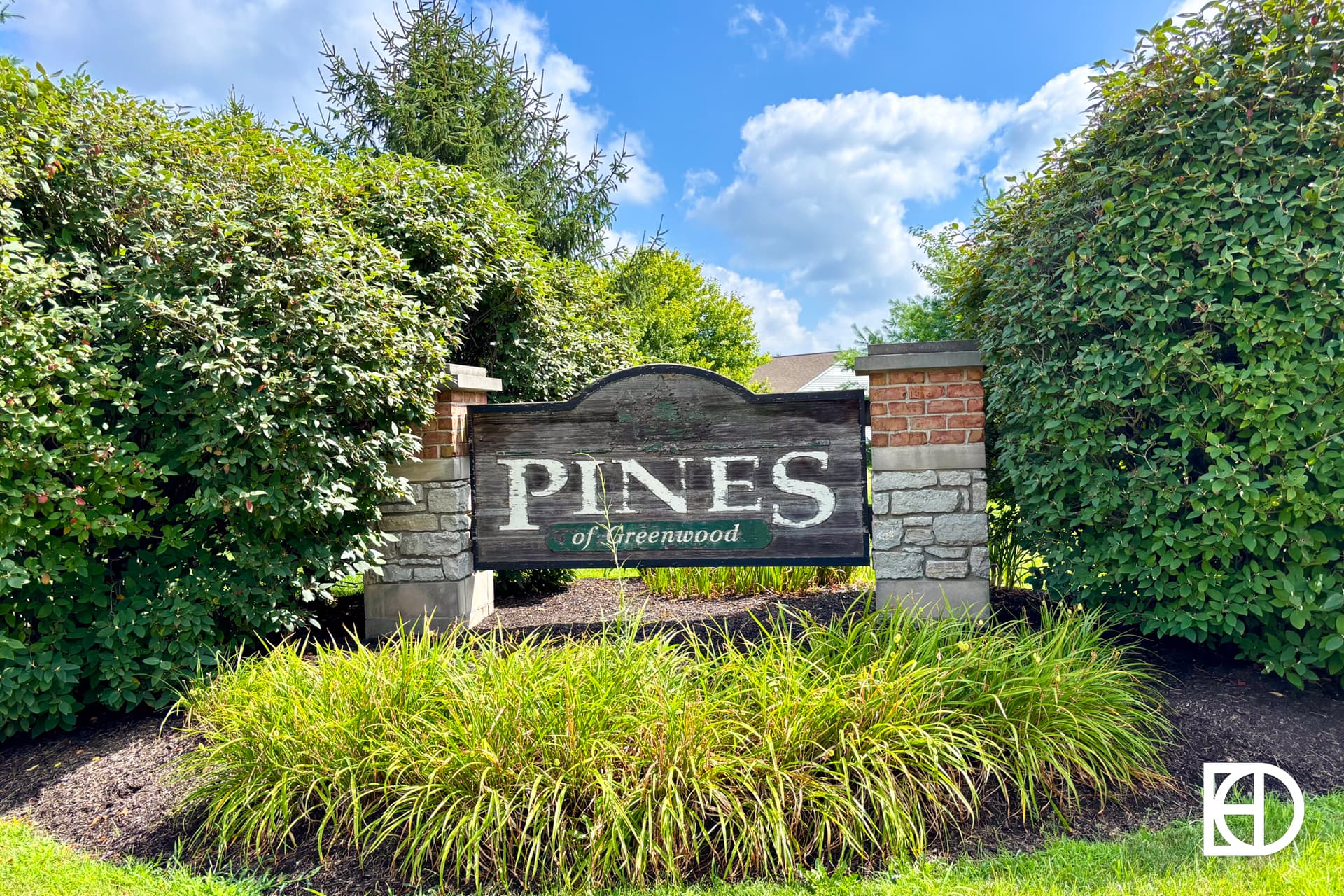 A wooden sign reading PINES of Greenwood is surrounded by green bushes, stone pillars, and bright green grass under a blue sky with clouds.