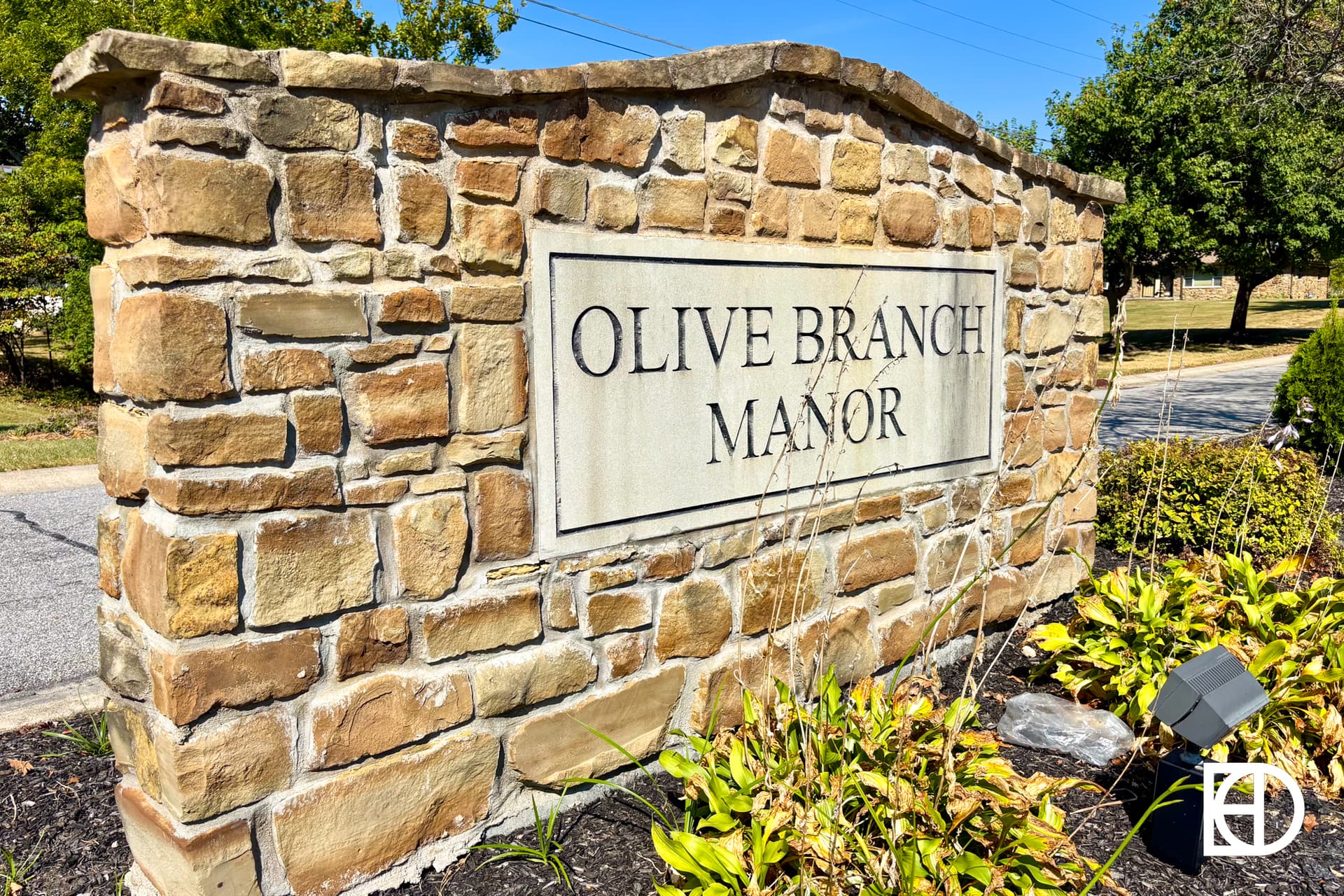 A stone sign with the words Olive Branch Manor is surrounded by greenery and landscaping, located next to a street on a sunny day.