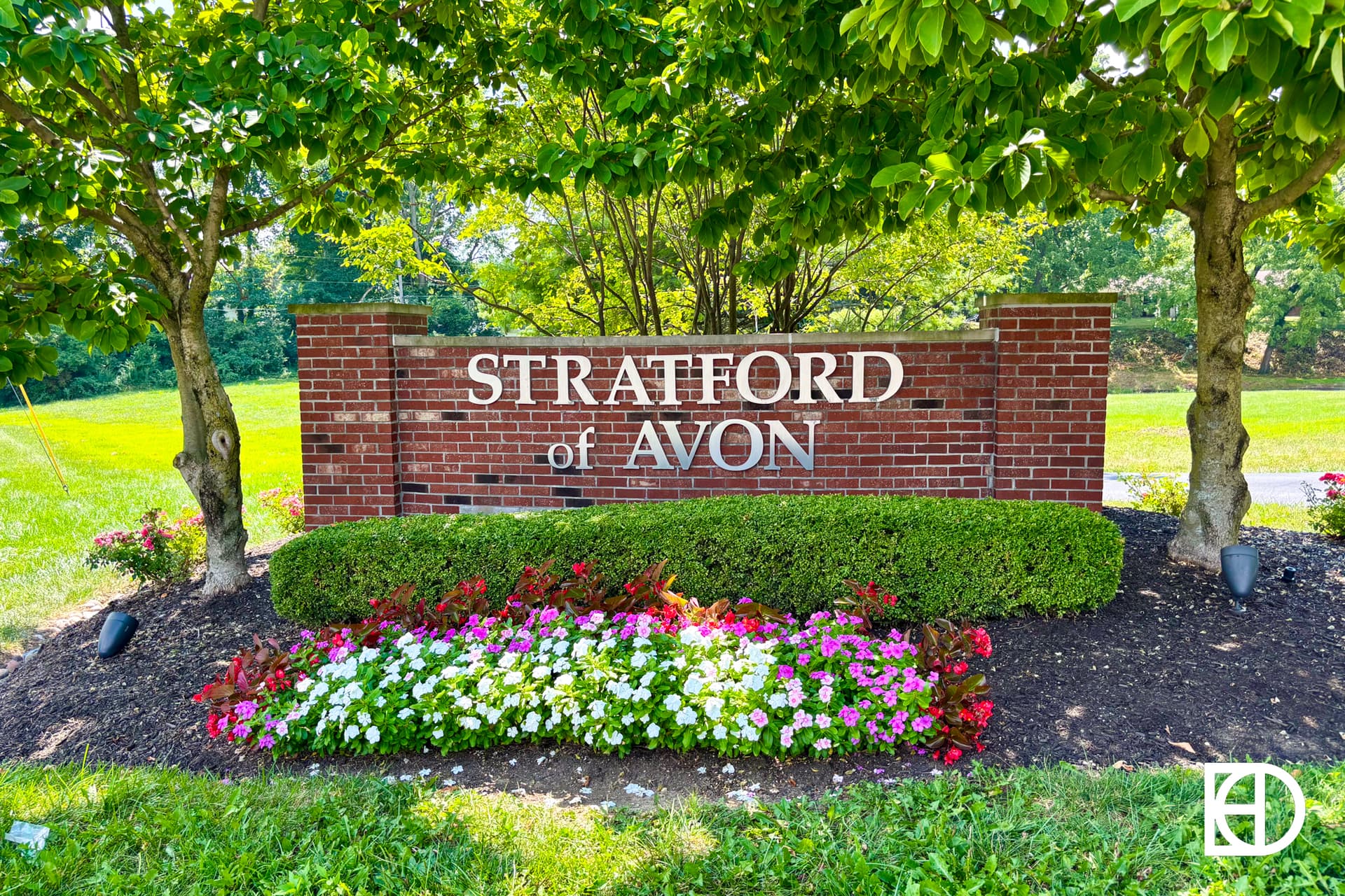 A brick sign reading Stratford of Avon stands between two trees, with neatly trimmed bushes and colorful flower beds in front, in a grassy, sunlit area.