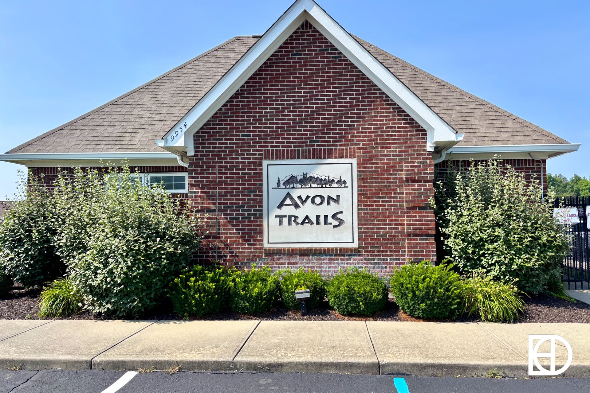 A red-brick building with a triangular roof features a large sign reading Avon Trails surrounded by bushes and landscaping, set against a clear blue sky.
