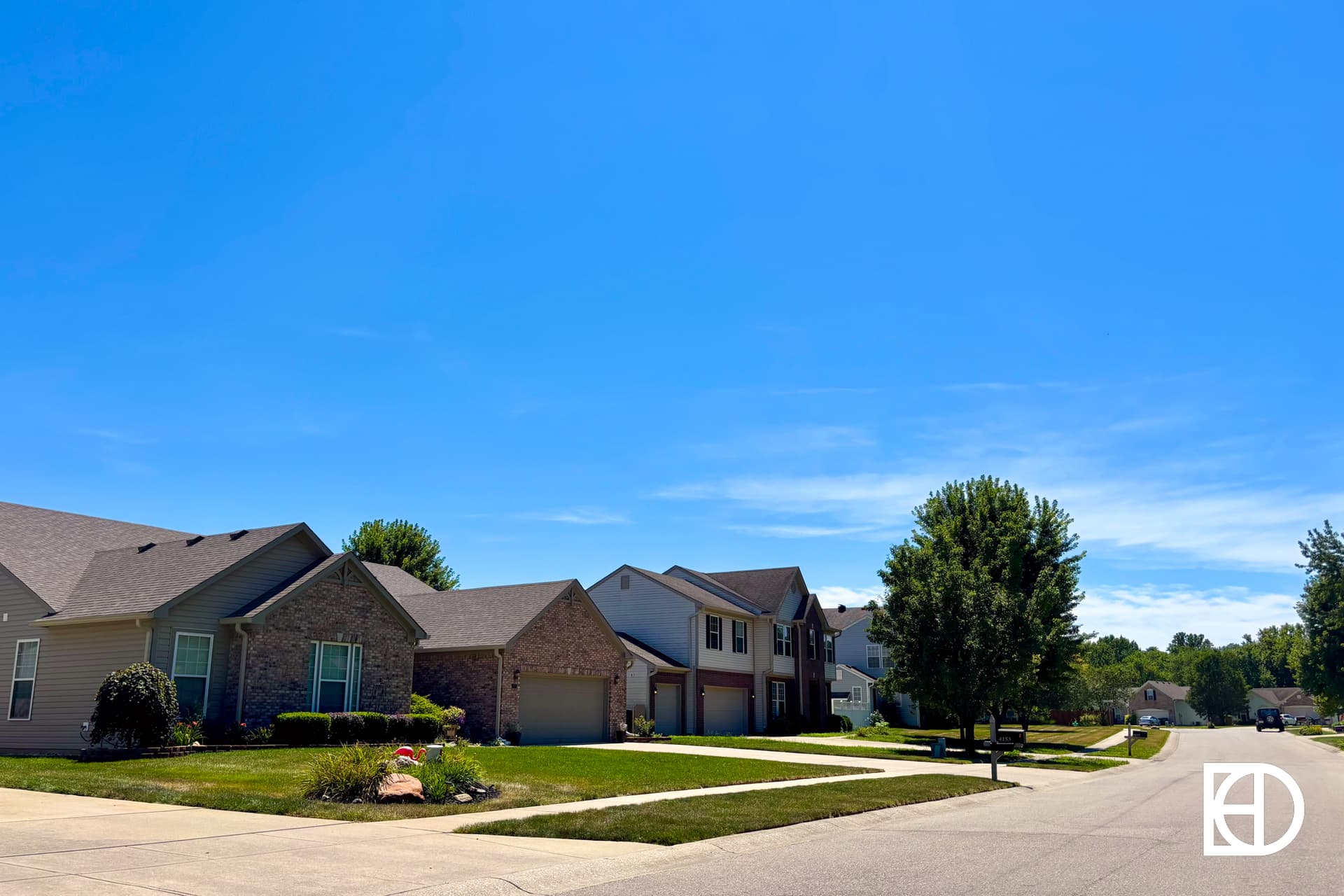 A suburban neighborhood with neatly kept lawns, modern houses, and a clear blue sky on a sunny day. A quiet street curves through the scene, with trees lining the road. A white logo is in the bottom right corner.