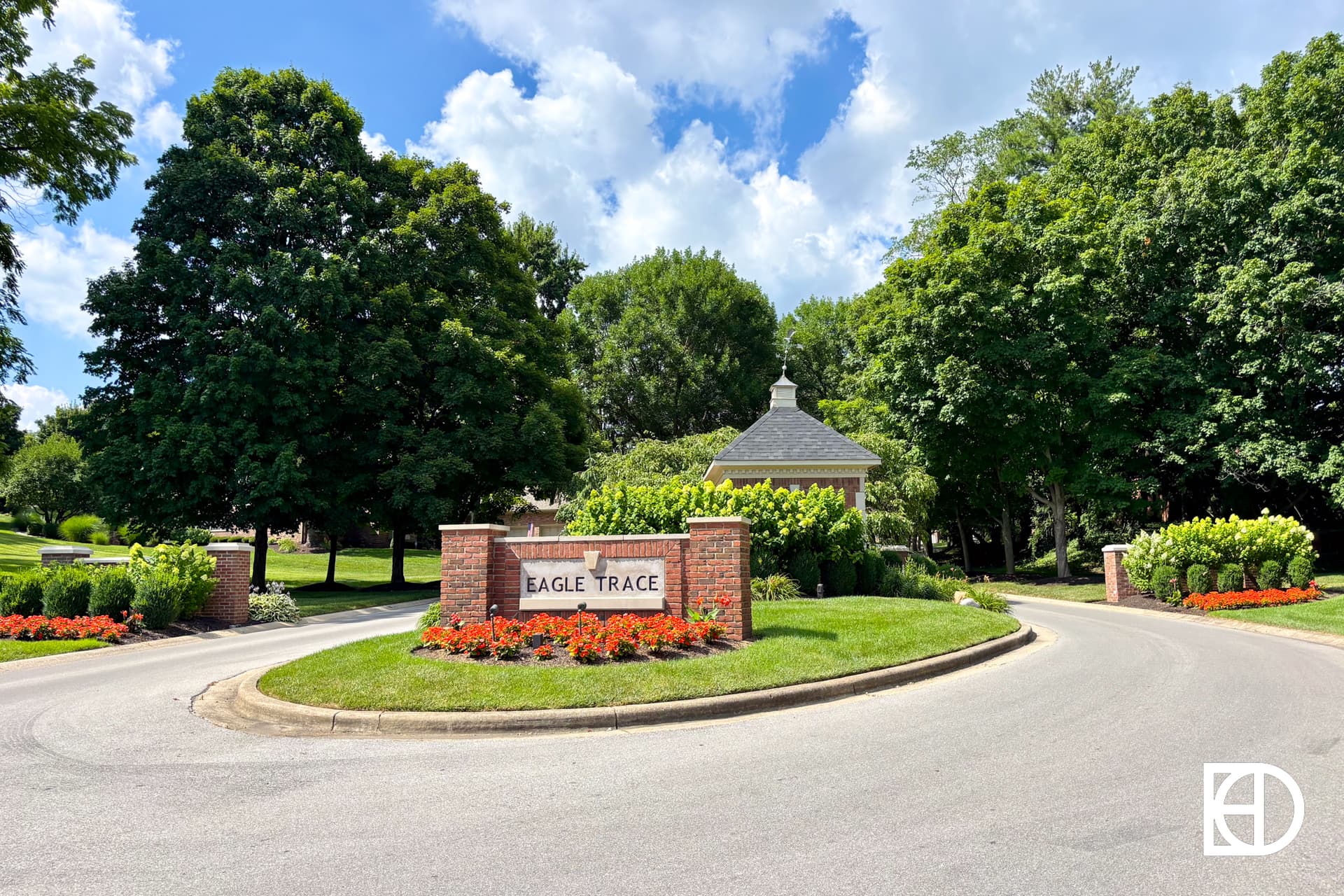 A brick entrance sign reading Eagle Trace sits between two pillars, surrounded by green lawns, bright flower beds, and tall trees under a partly cloudy blue sky. A small building is visible behind the sign.