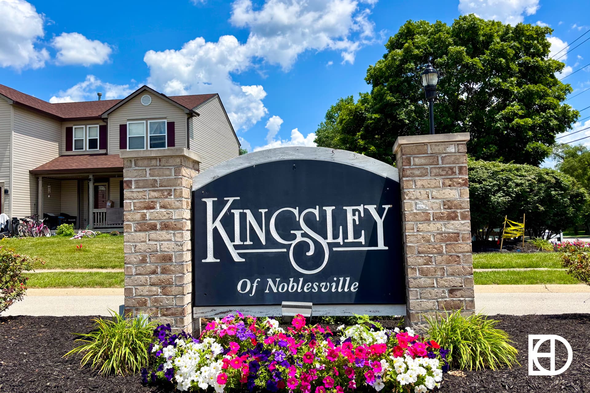A stone and metal sign reads Kingsley of Noblesville in front of a house, with colorful flowers planted at the base and trees and a blue sky in the background.