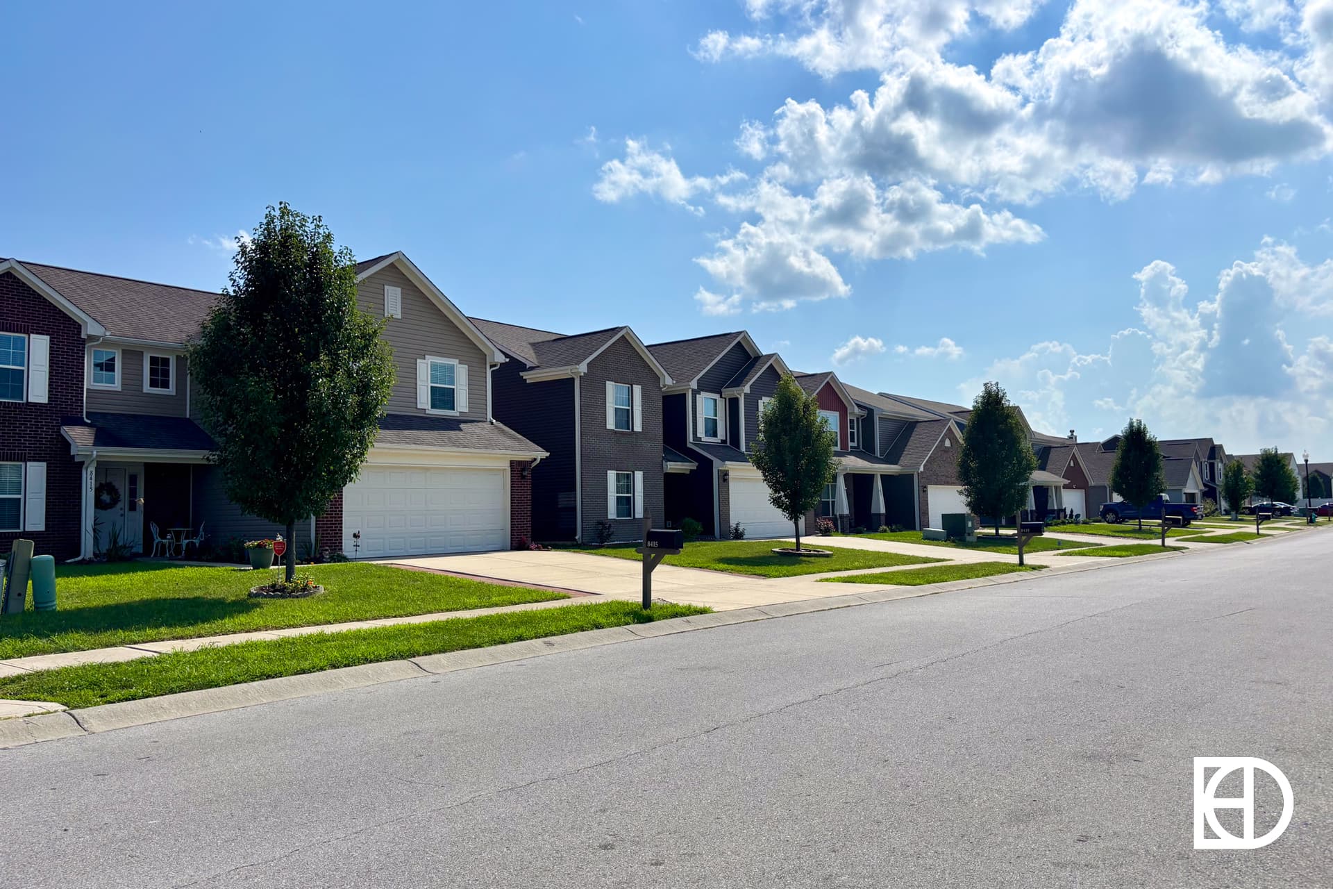 A sunny suburban street with rows of two-story houses, each with driveways, garages, and small front lawns. Trees line the street and a few clouds are visible in the blue sky.