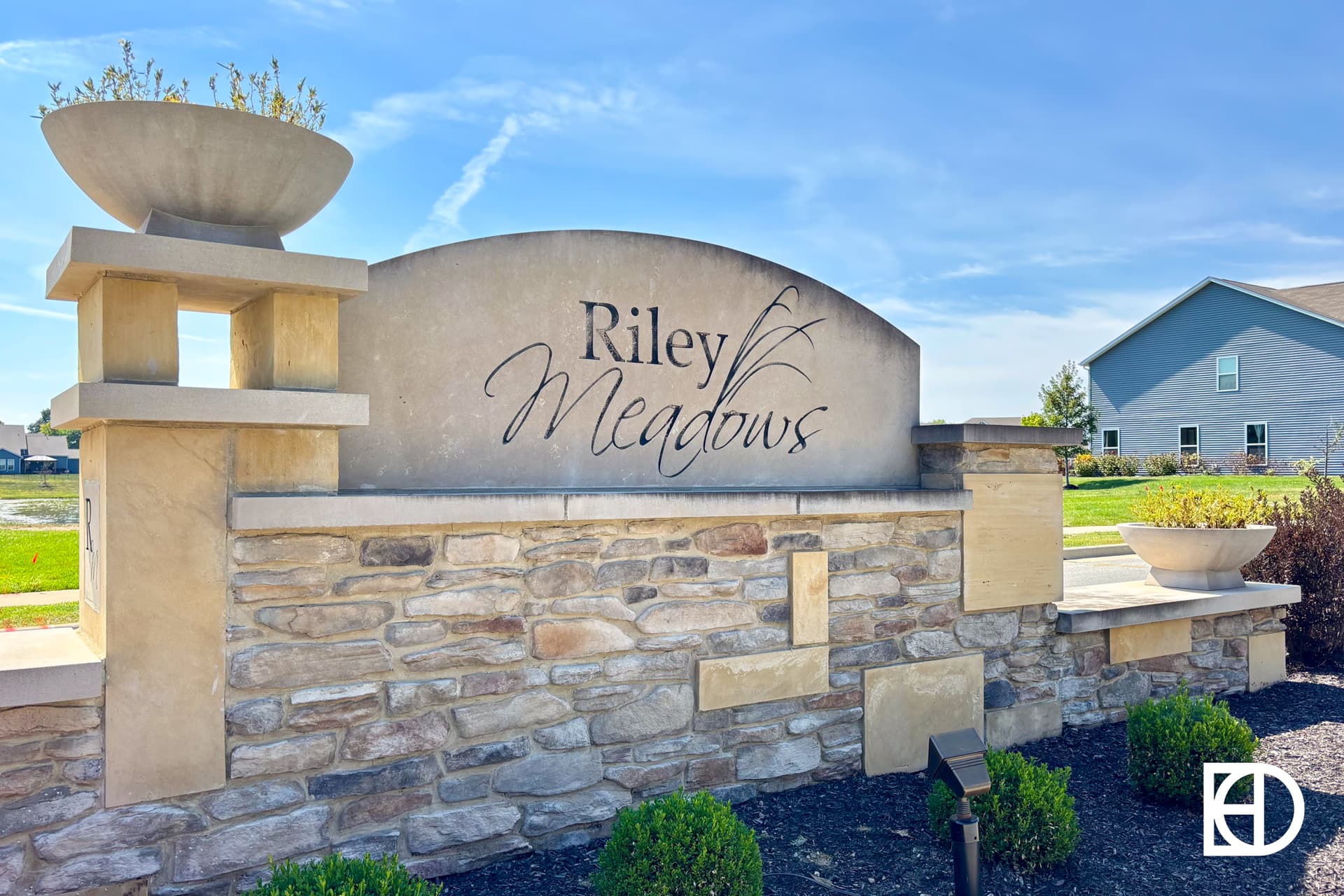 Stone entrance sign for Riley Meadows with decorative planters on each side, surrounded by landscaping and a modern house visible in the background under a clear blue sky.