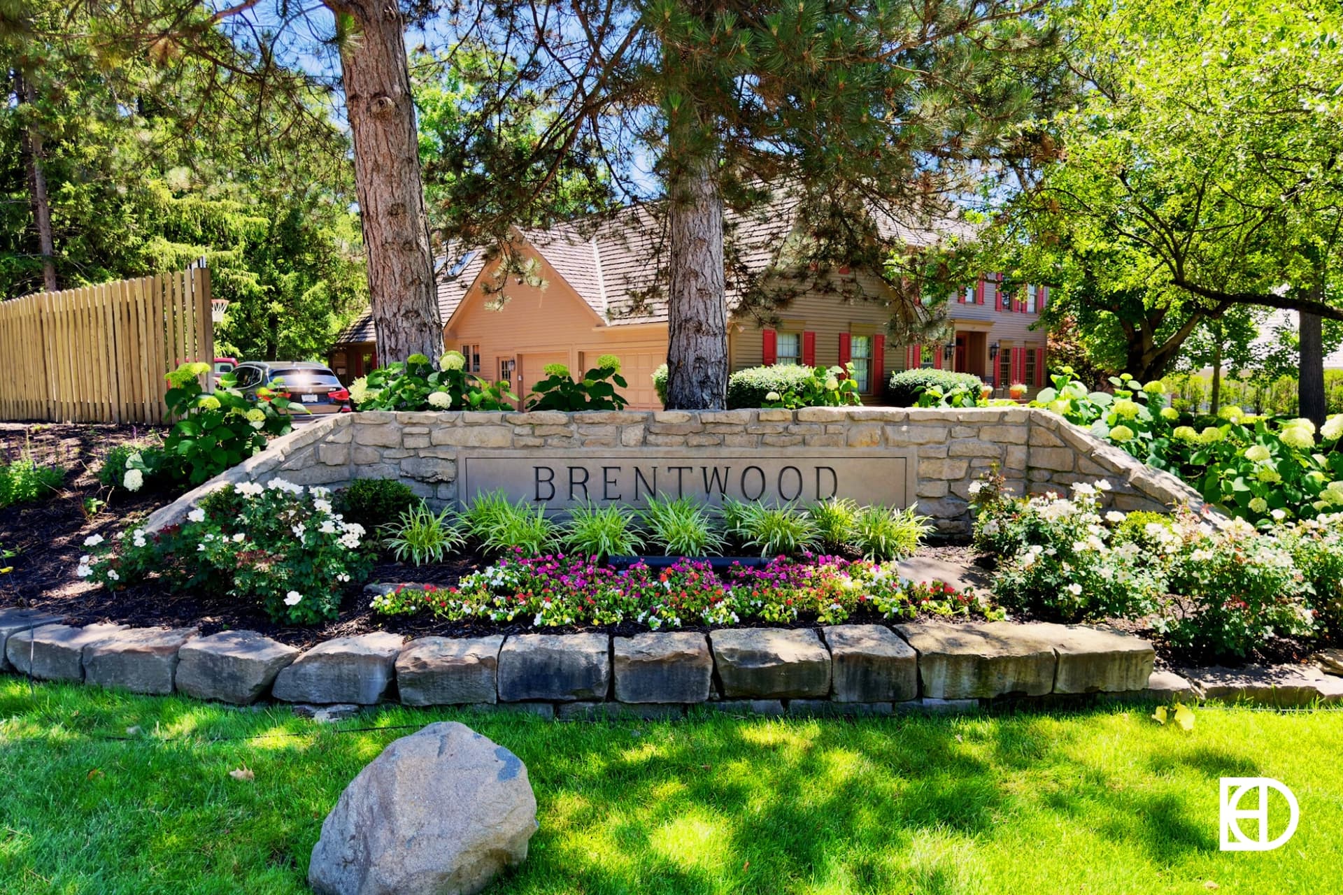Stone entrance sign for Brentwood featuring large paver edging around flower bed with shrubs and flowers, and tall trees behind.