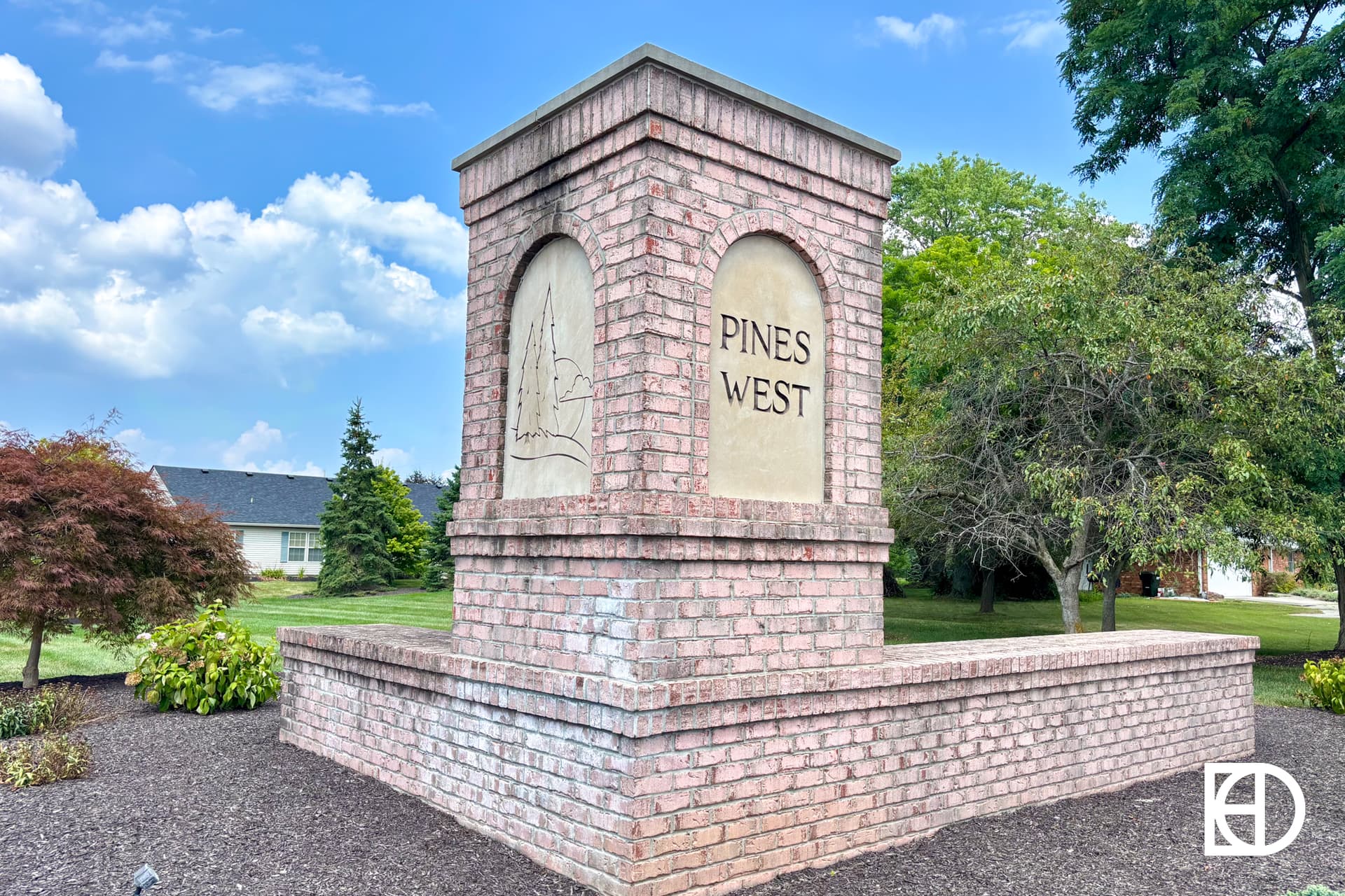 A brick monument sign with “PINES WEST” engraved on it stands surrounded by mulch, greenery, and trees under a blue sky with scattered clouds.