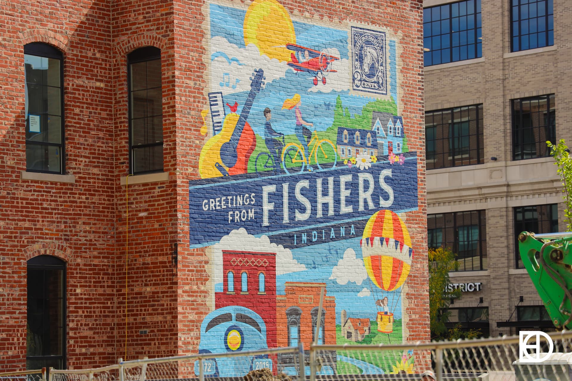 A colorful mural on a brick building reads Greetings from Fishers Indiana with illustrations of local landmarks, a guitar, bicycles, and a hot air balloon.