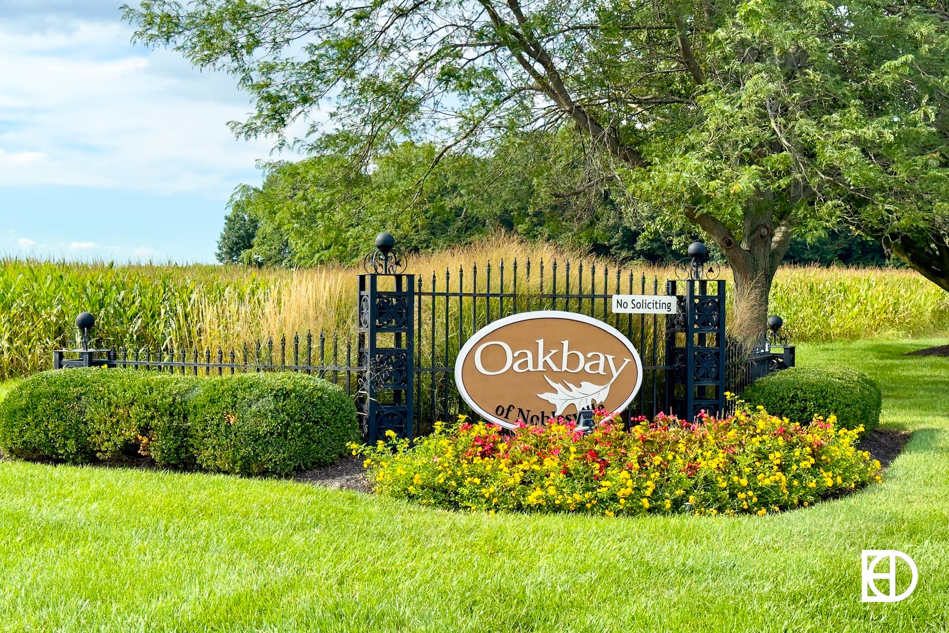 Oval entrance sign set on iron gate with landscaping surround and corn field in background.