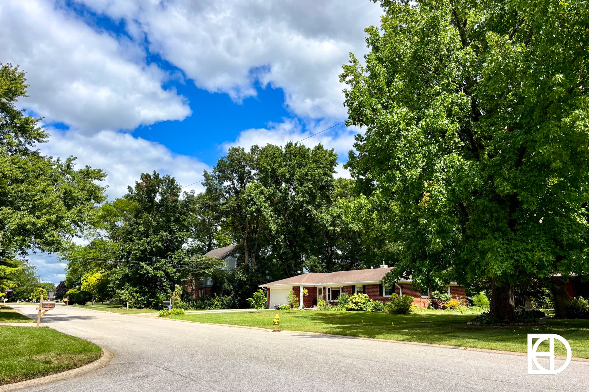 A suburban street curves past green lawns, large leafy trees, and single-story brick homes under a partly cloudy blue sky.