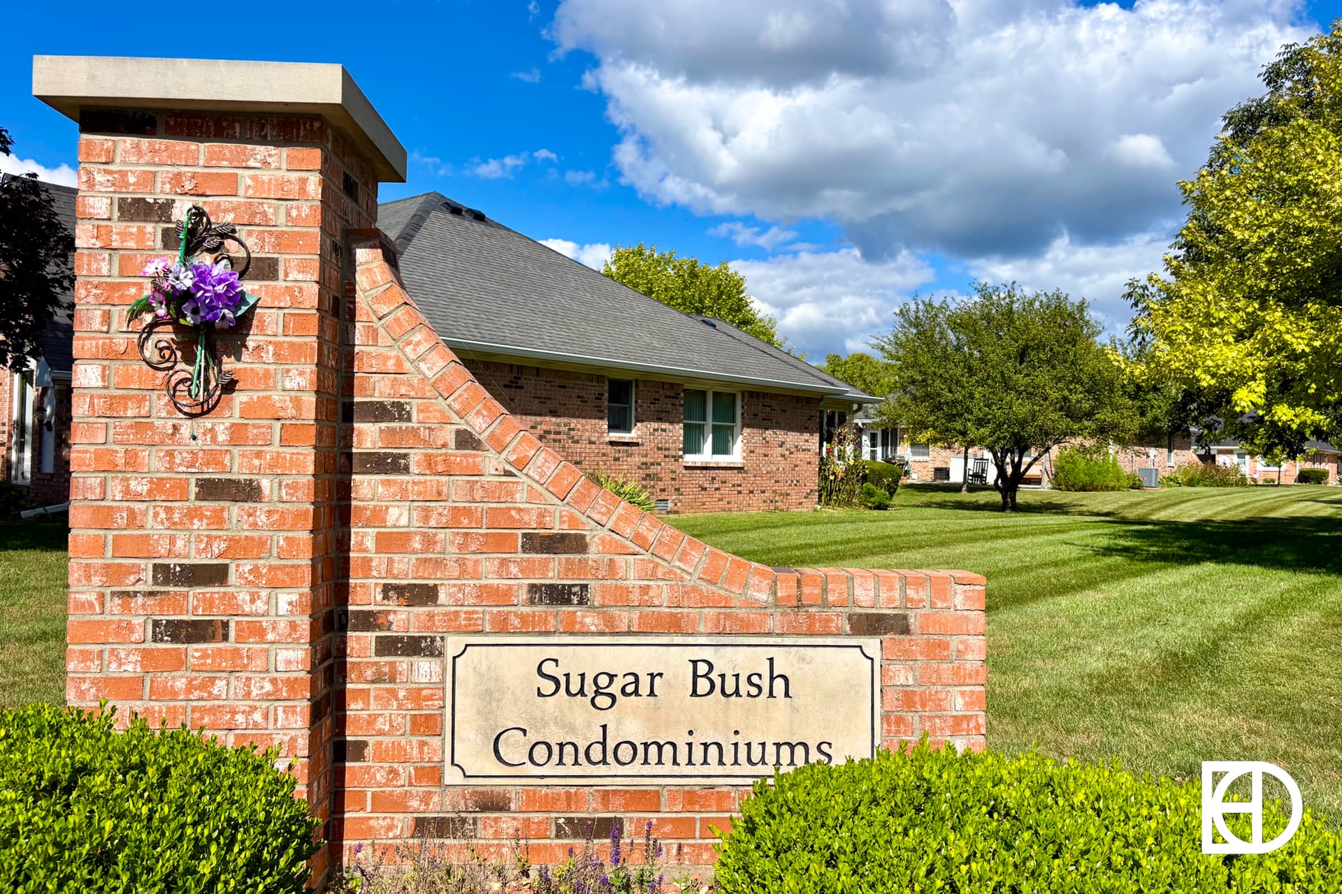 A brick sign reading “Sugar Bush Condominiums” stands on a well-kept lawn, with residential buildings and trees in the background under a bright blue sky with scattered clouds.