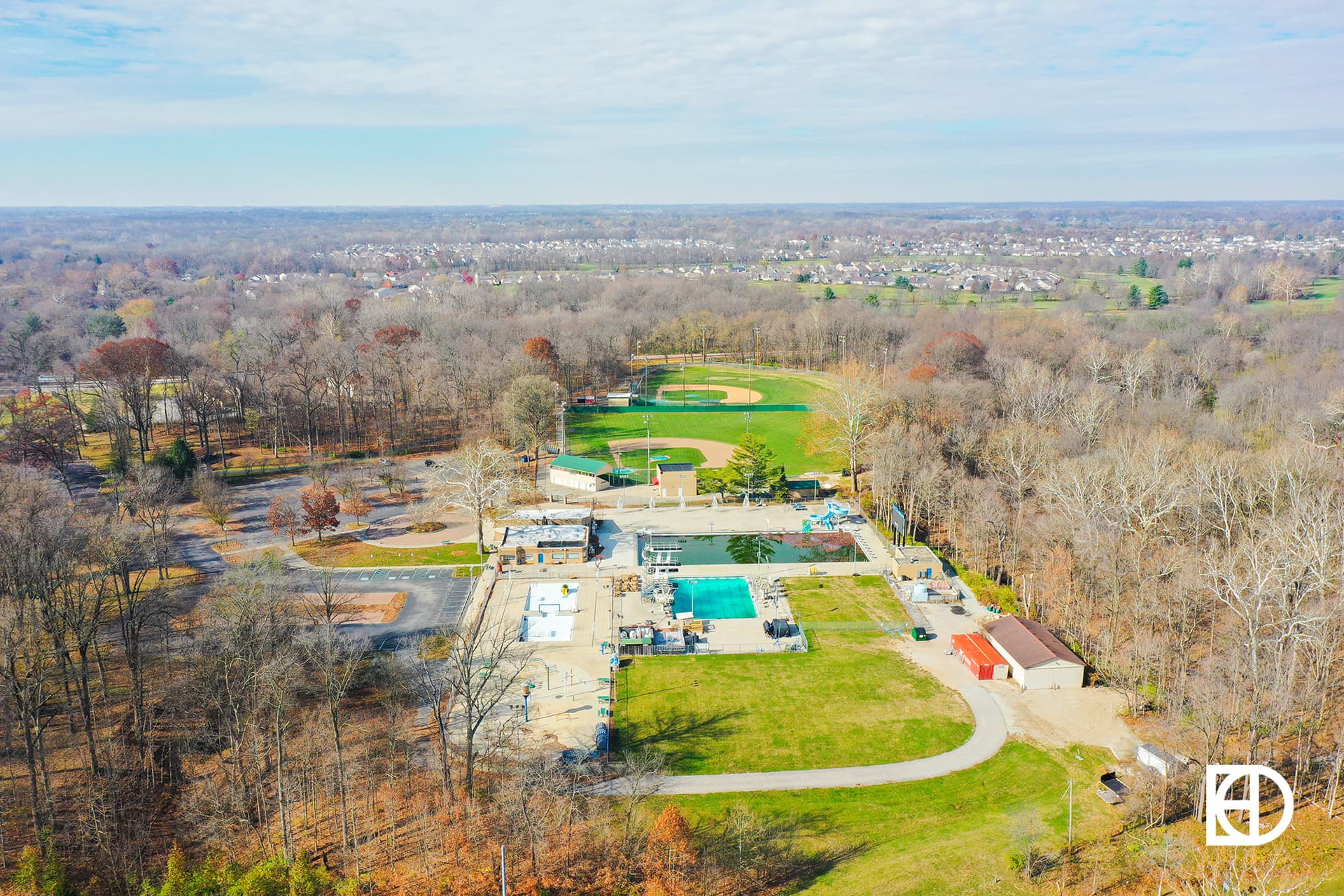 Aerial view of a park with a swimming pool, baseball field, parking lot, and surrounding trees during autumn.