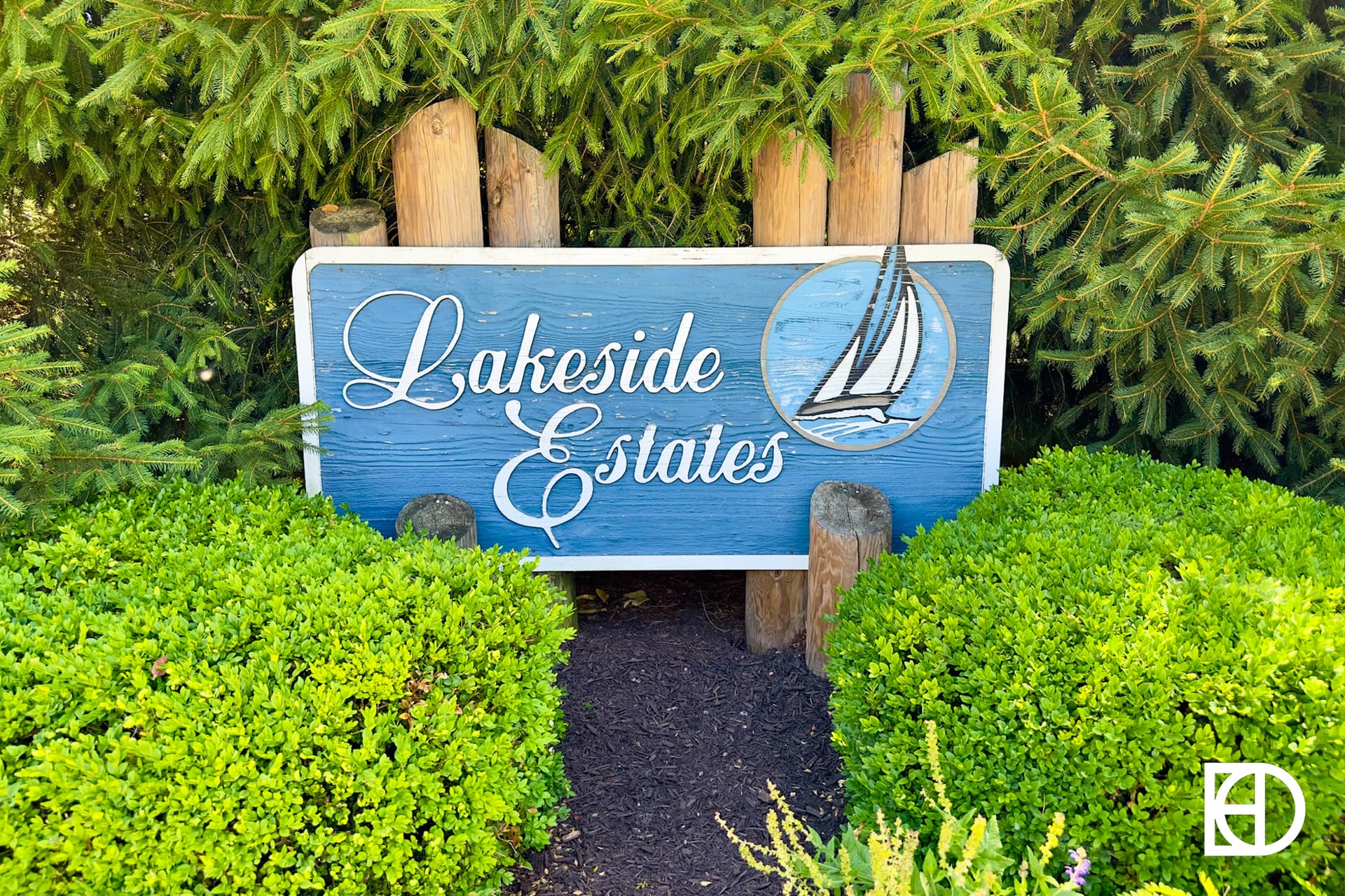 A blue wooden sign with white text reads “Lakeside Estates” and features an image of a sailboat. The sign is surrounded by green bushes and pine trees.