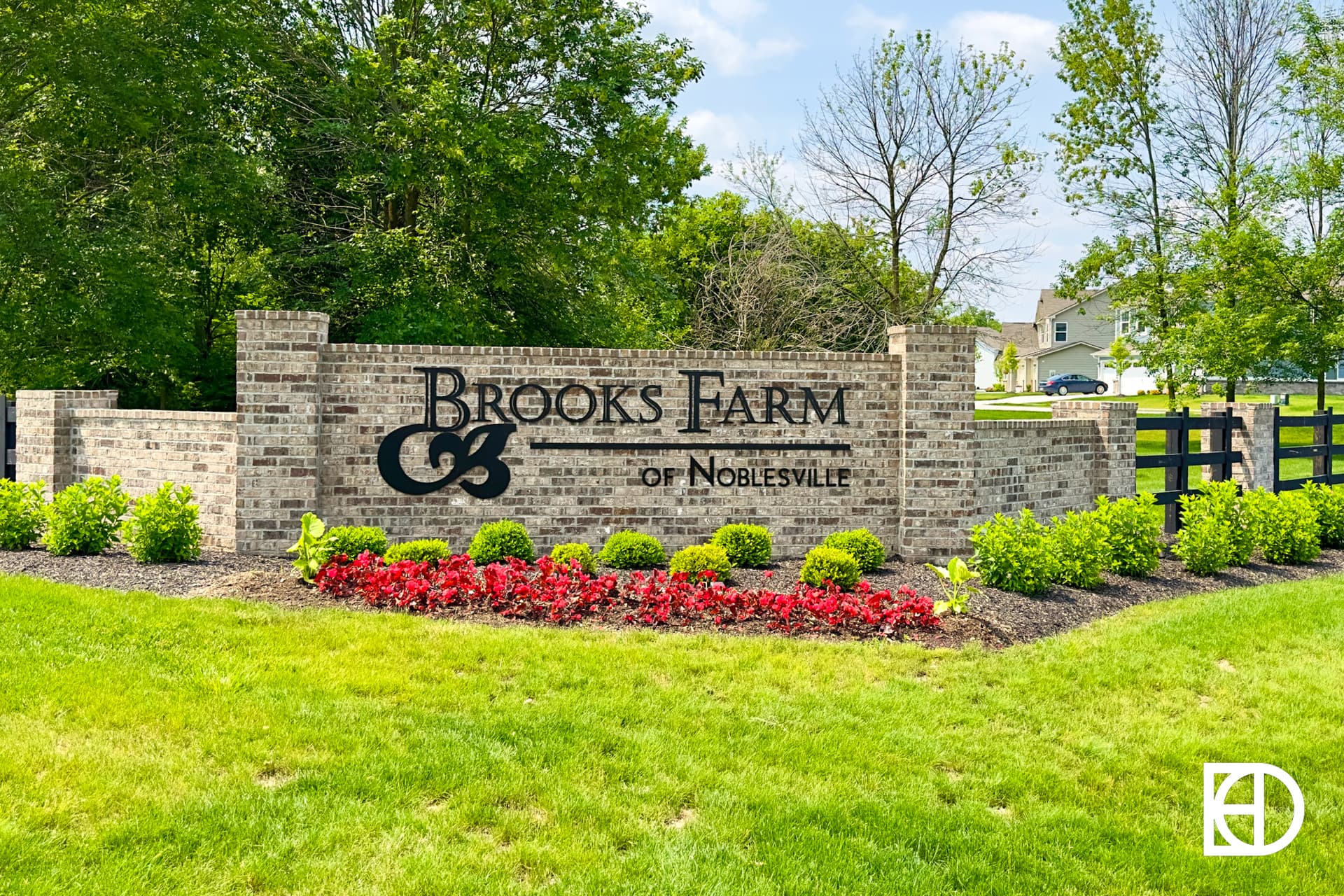 A wide grassy field with power lines running diagonally across a clear blue sky. In the background are houses, trees, and a white fence.