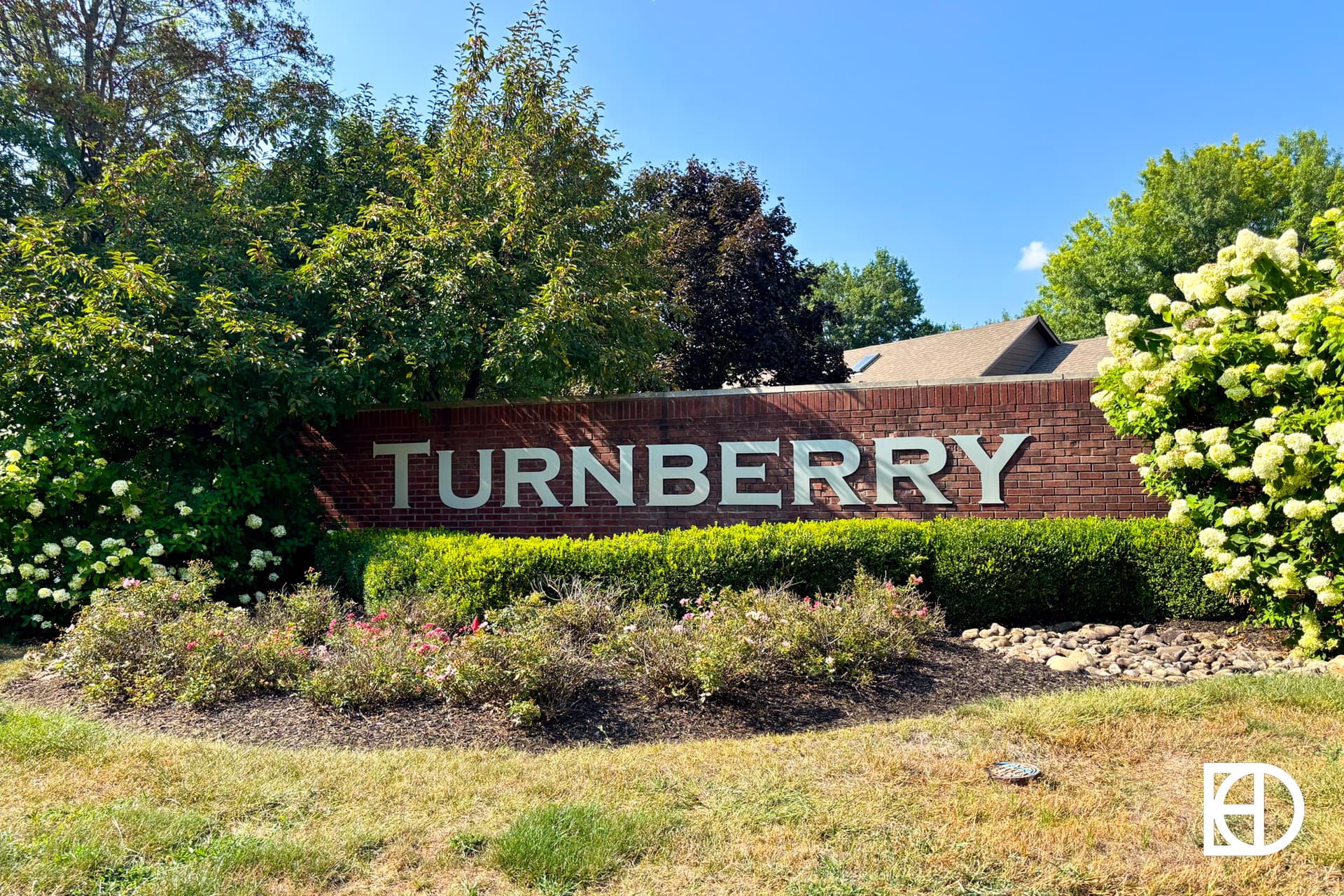 A brick sign reads TURNBERRY in large silver letters, surrounded by green bushes, flowering plants, and trees under a clear blue sky.