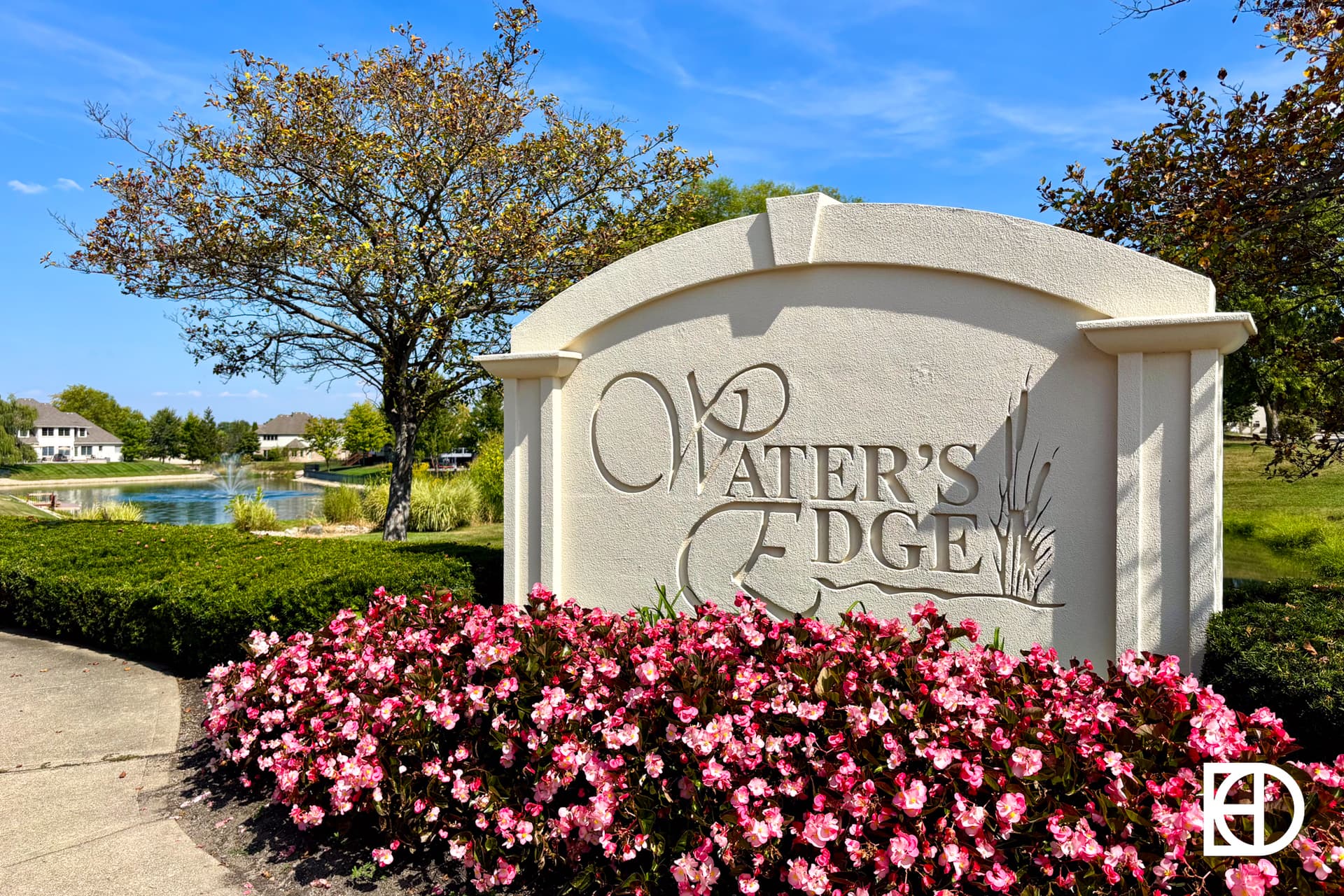 A large white sign reading “Water’s Edge” stands in front of pink flowers and manicured bushes, with a pond, houses, and trees visible in the background on a sunny day.
