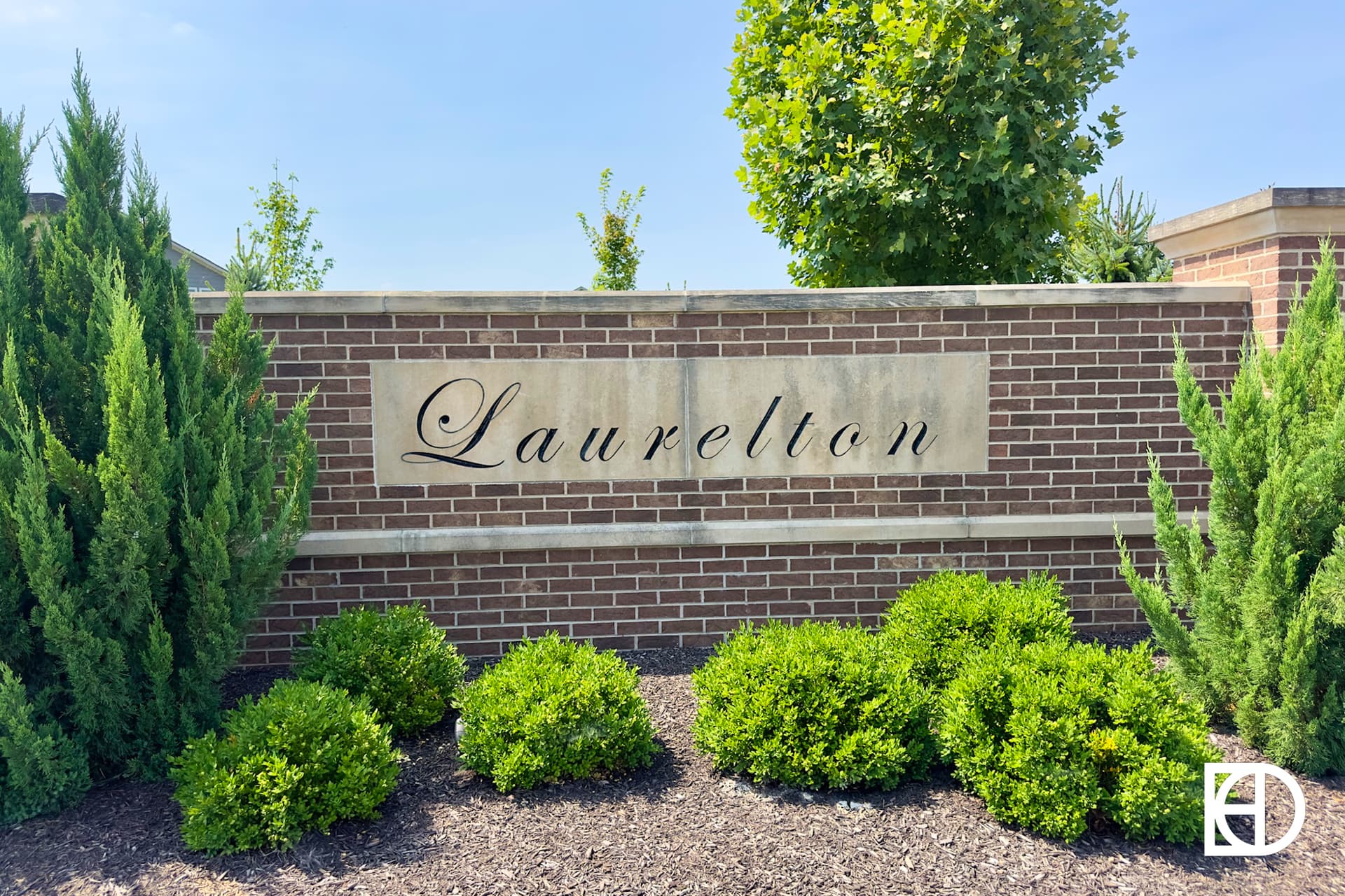 A brick entrance sign with the word Laurelton in script letters, surrounded by green shrubs and with a large leafy tree in the background.