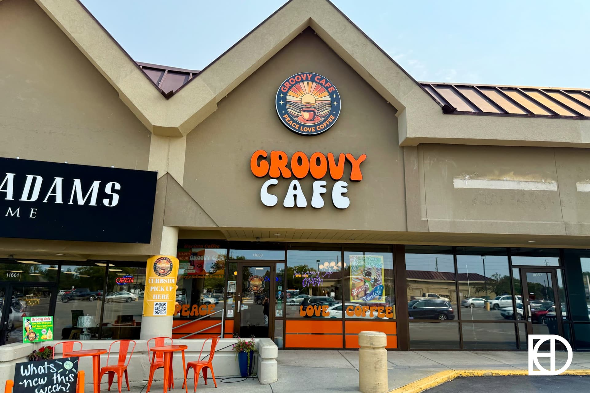 Exterior of Groovy Cafe with orange sign, round logo above, large windows, and two orange tables with chairs outside. The entrance features peace-themed décor and a sign that says “LOVE COFFEE.”.