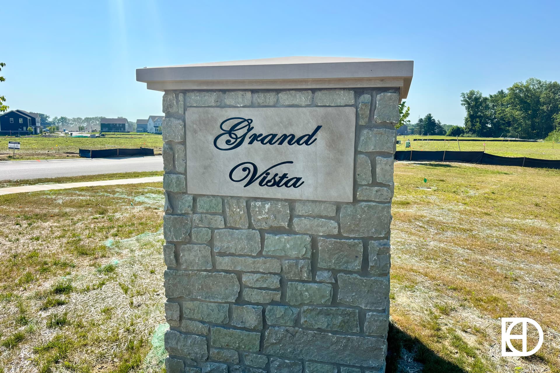 A stone monument sign with Grand Vista written in script stands on a grassy lot in a suburban neighborhood under a clear blue sky.