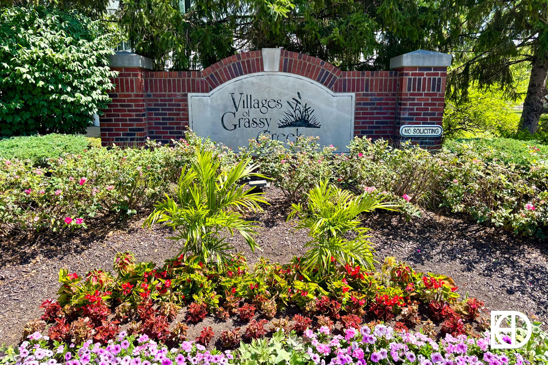 A brick and stone entrance sign reads Villages at Grassy Creek,” surrounded by green shrubs, colorful flowers, and trees. A small sign nearby says “No Soliciting.”.