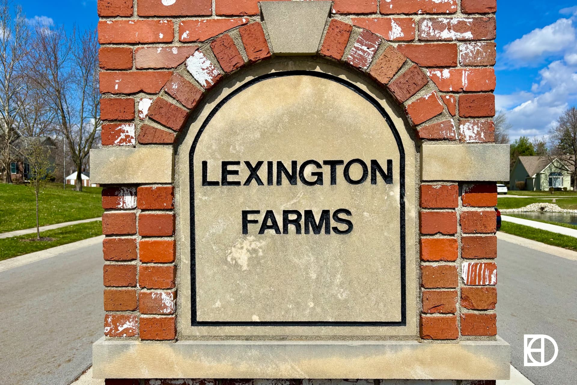 A stone and brick sign reads Lexington Farms at the entrance to a residential neighborhood, with houses and trees visible in the background.
