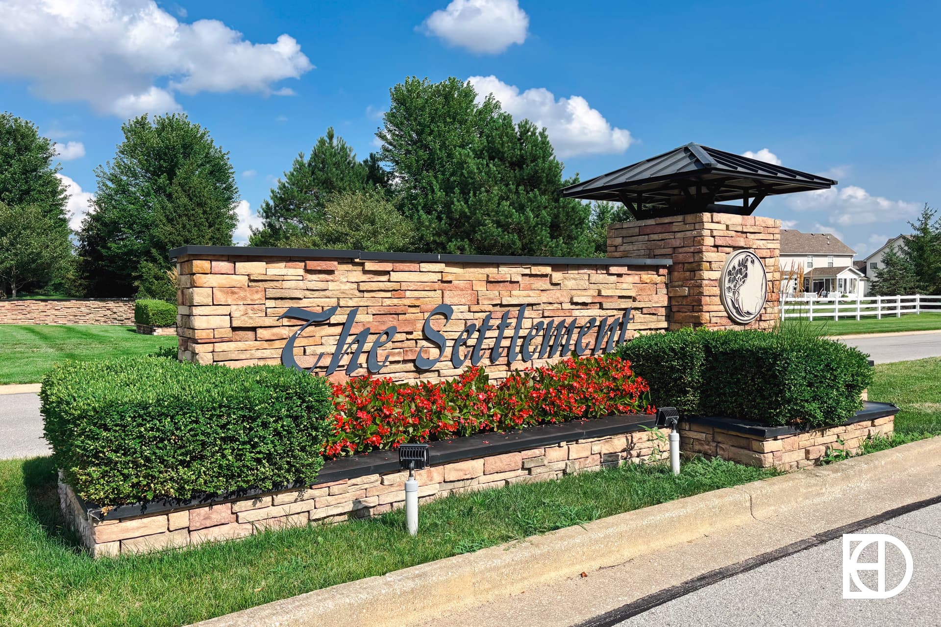 A stone entrance sign with The Sutherlands in large letters, surrounded by green bushes and red flowers, stands near a road with trees, grass, and houses in the background under a blue sky.