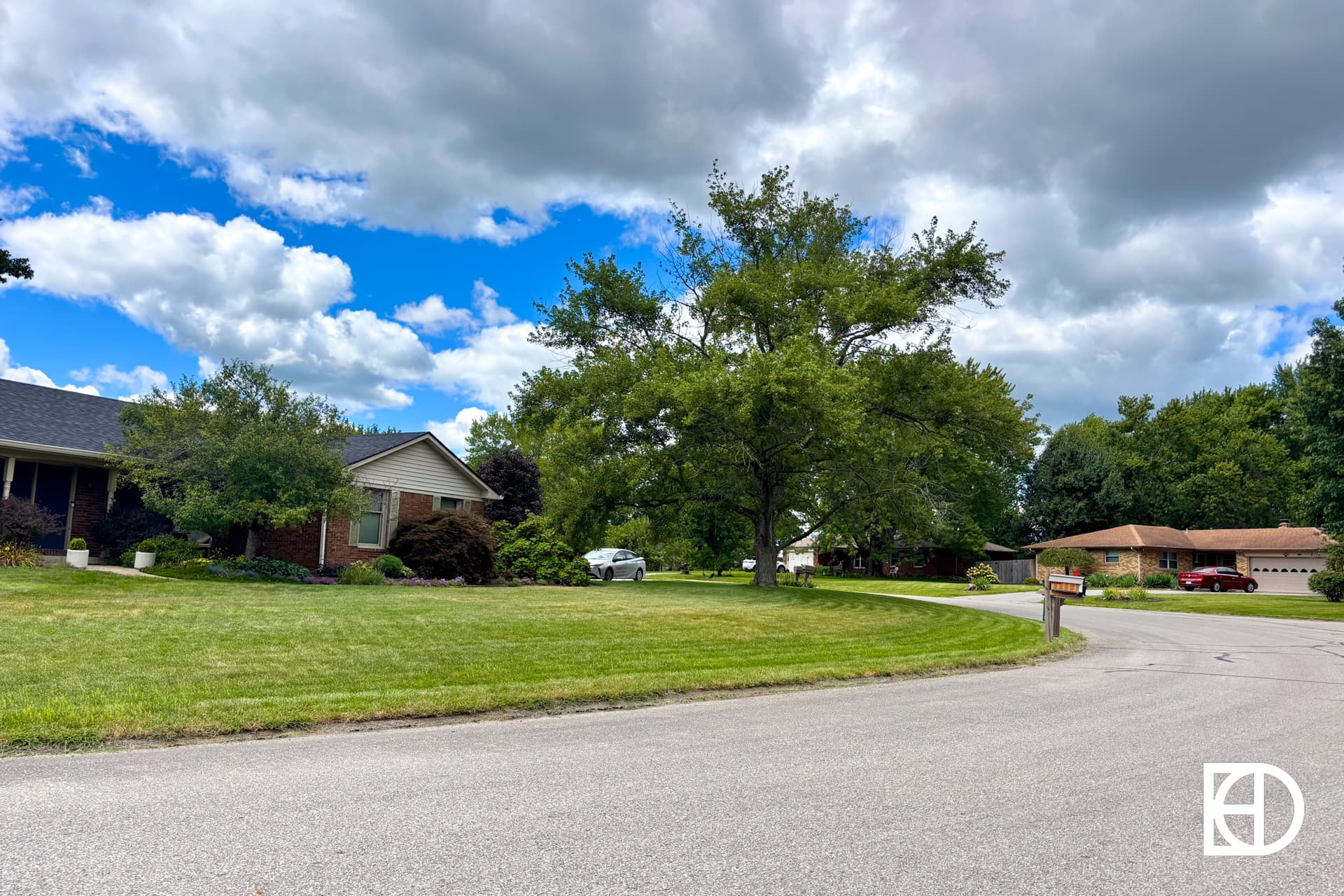A suburban neighborhood with houses, green lawns, large trees, and a curving street under a partly cloudy sky. A white car is parked in a driveway. The KD logo is in the bottom right corner.