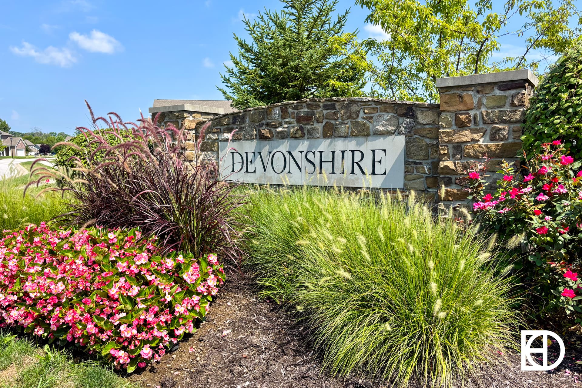 A stone entrance sign with DEVONSHIRE written on it, surrounded by colorful flowers, ornamental grasses, and greenery under a blue sky.