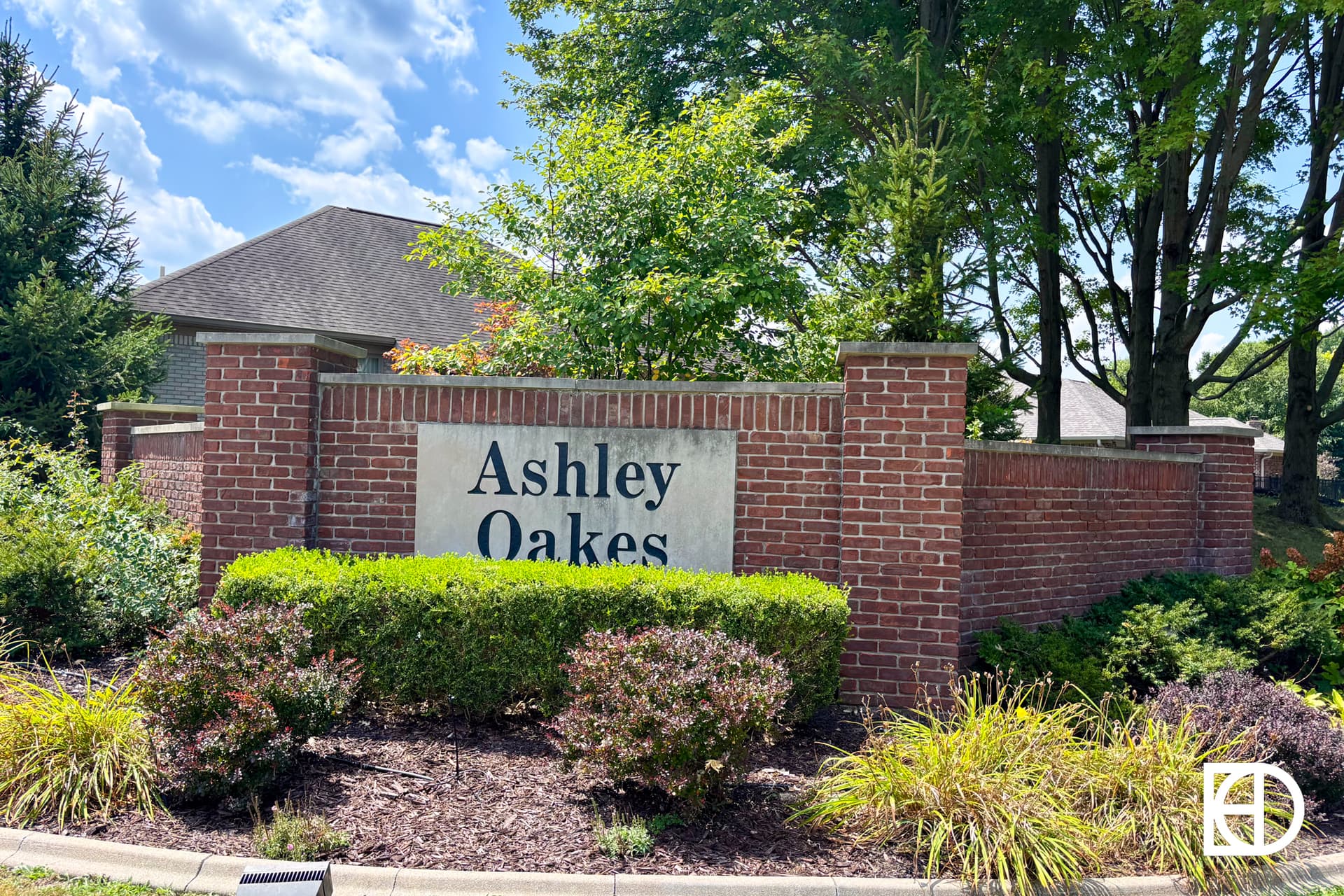 A brick sign with “Ashley Oakes” in black letters is surrounded by bushes and trees. A house with a sloped roof is partially visible behind the sign under a partly cloudy sky.