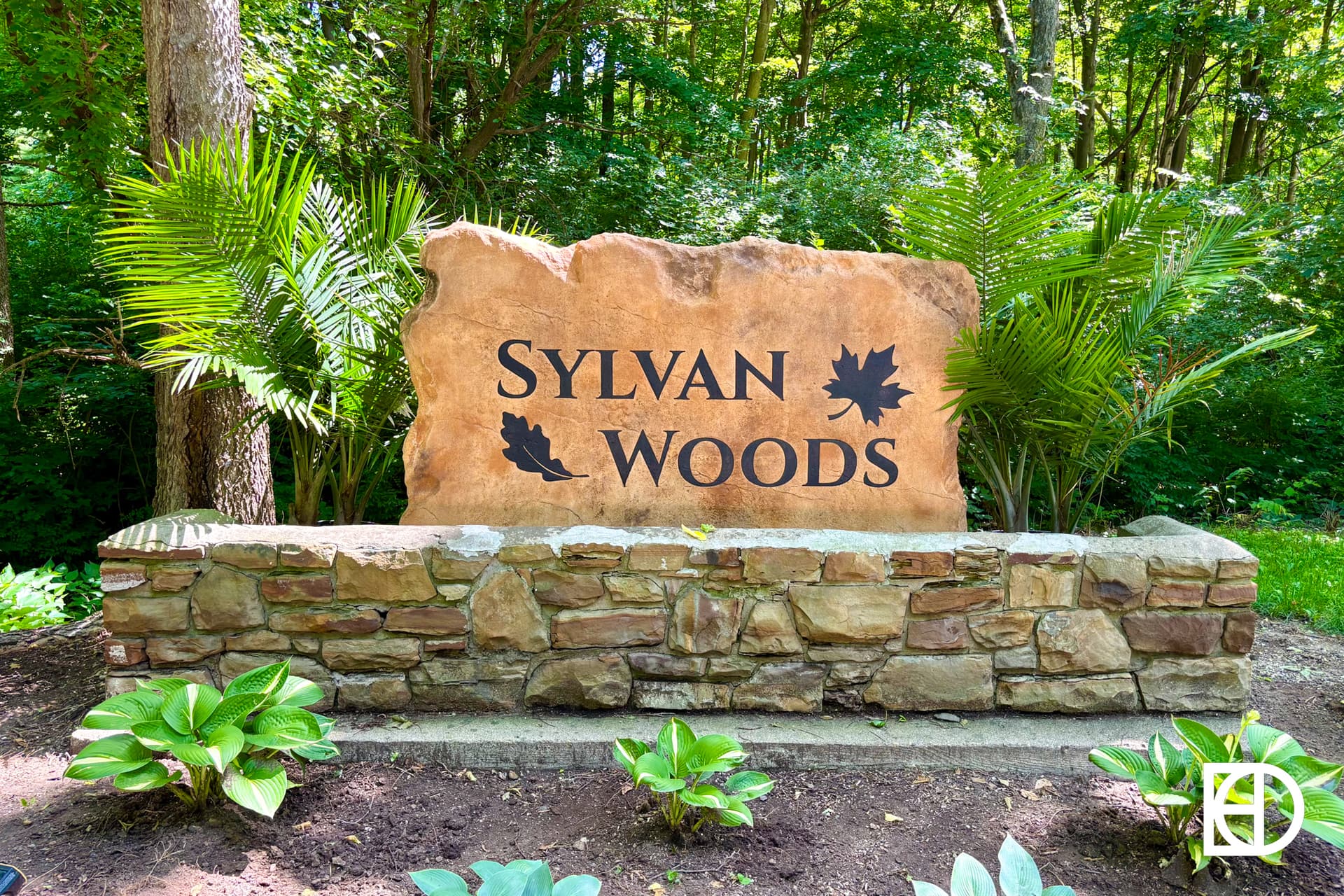 A large stone sign reading SYLVAN WOODS with two leaf graphics, surrounded by lush greenery and plants, stands in a wooded area with sunlight filtering through the trees.