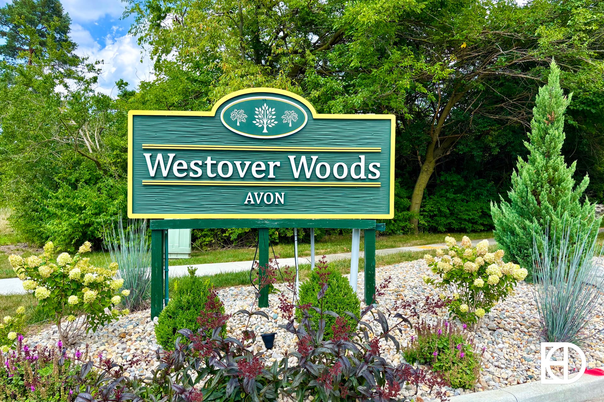 A green sign with yellow trim reads Westover Woods Avon surrounded by landscaped plants, small shrubs, and decorative rocks, with trees and greenery in the background.