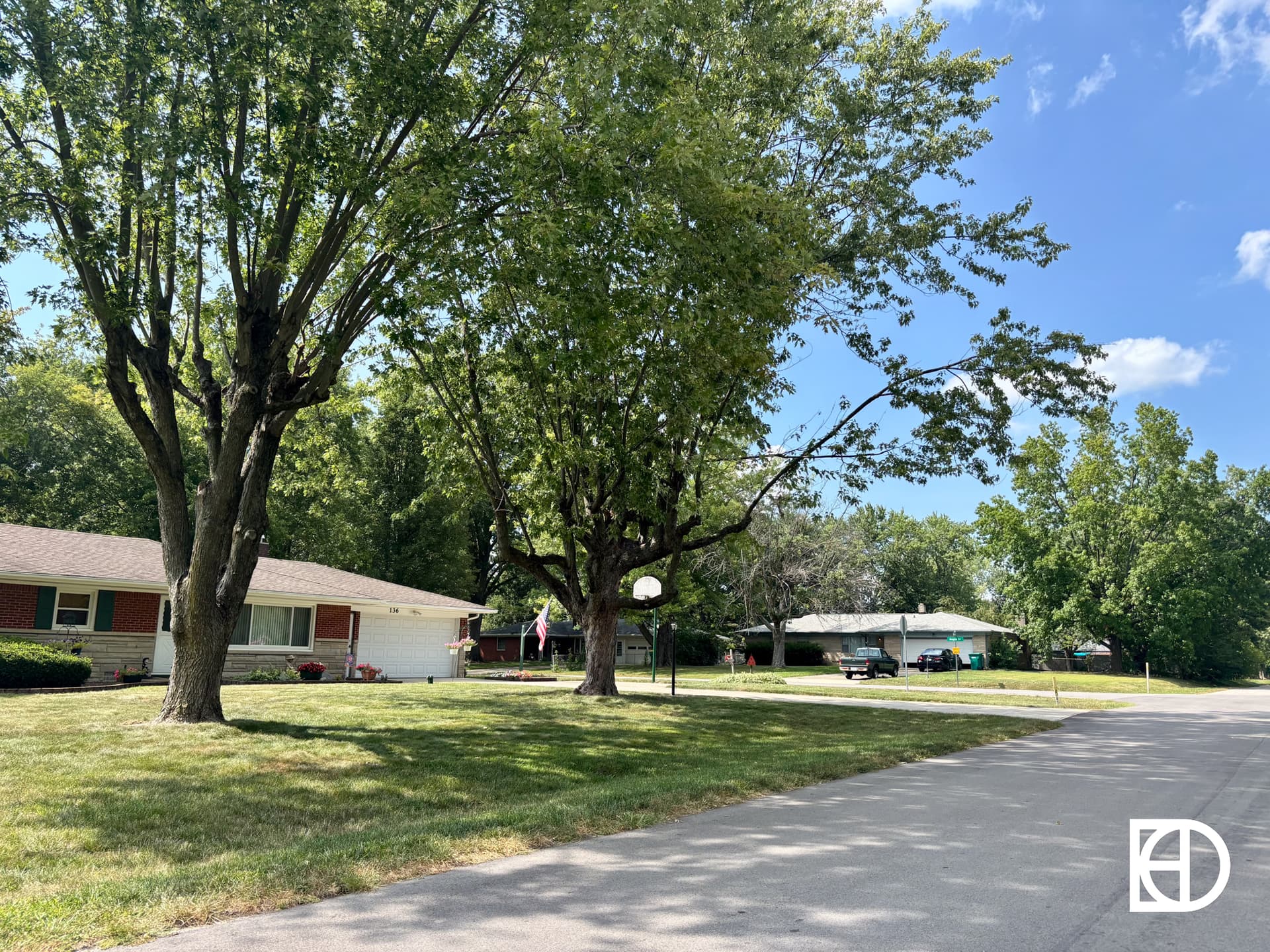 A quiet suburban street with large trees, green lawns, and single-story houses under a blue sky with scattered clouds. An American flag hangs by one house, and a basketball hoop stands near the sidewalk.