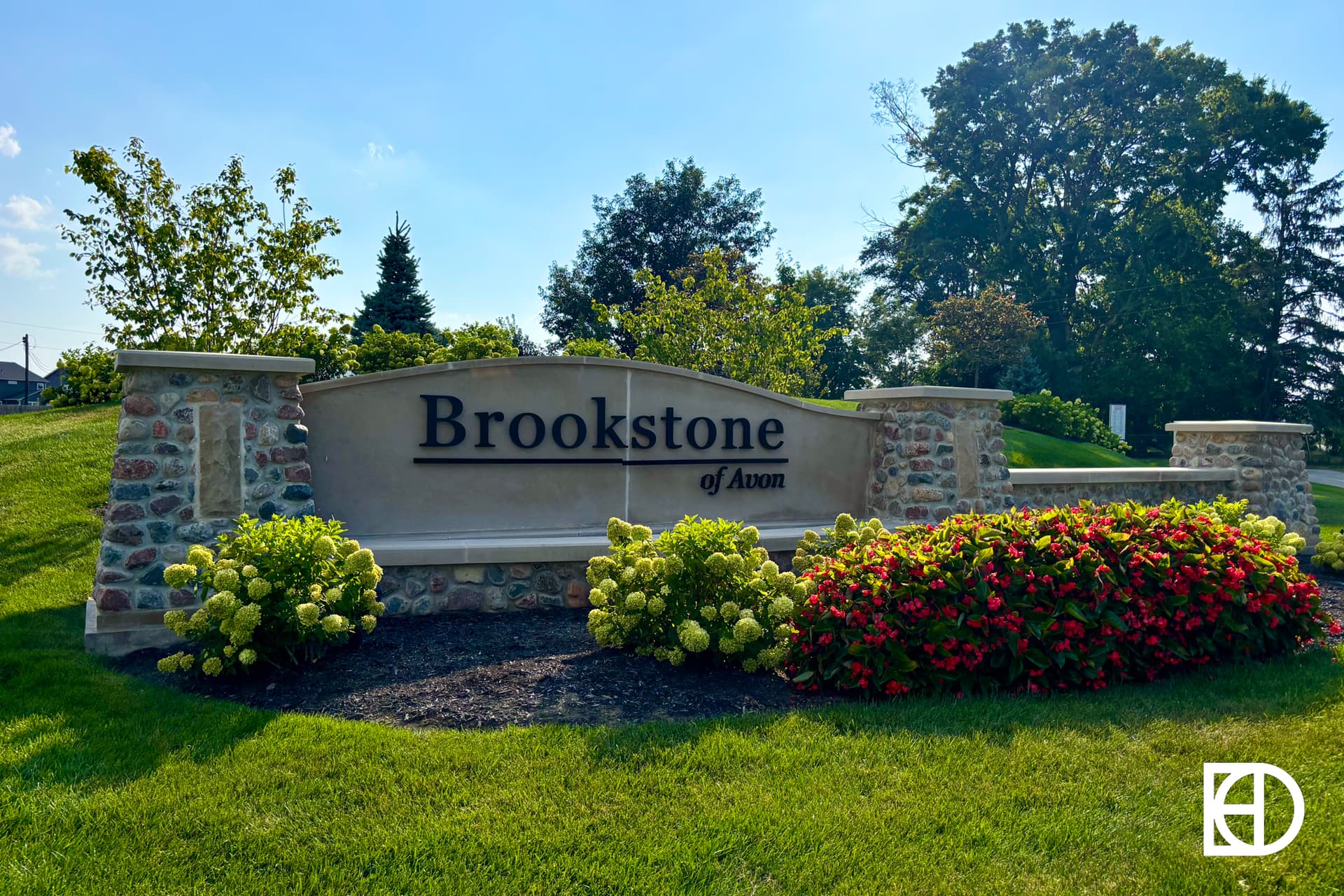 A stone entrance sign reads Brookstone of Avon, surrounded by green grass, blooming yellow and red flowers, and trees, on a sunny day. A white logo is in the lower right corner.