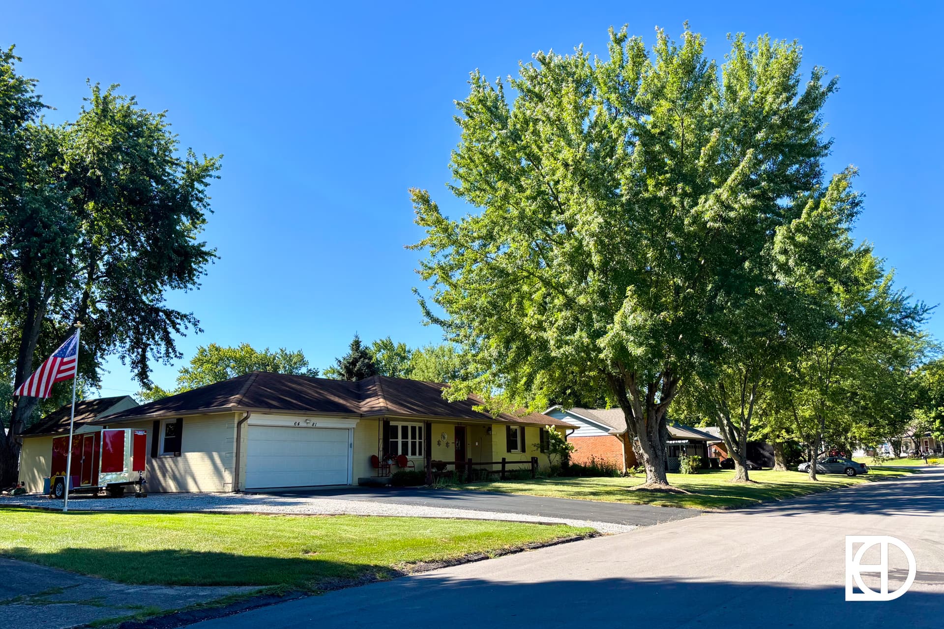 A quiet suburban street with single-story houses, large green trees lining the road, and an American flag on a pole in front of one home, all under a clear blue sky.