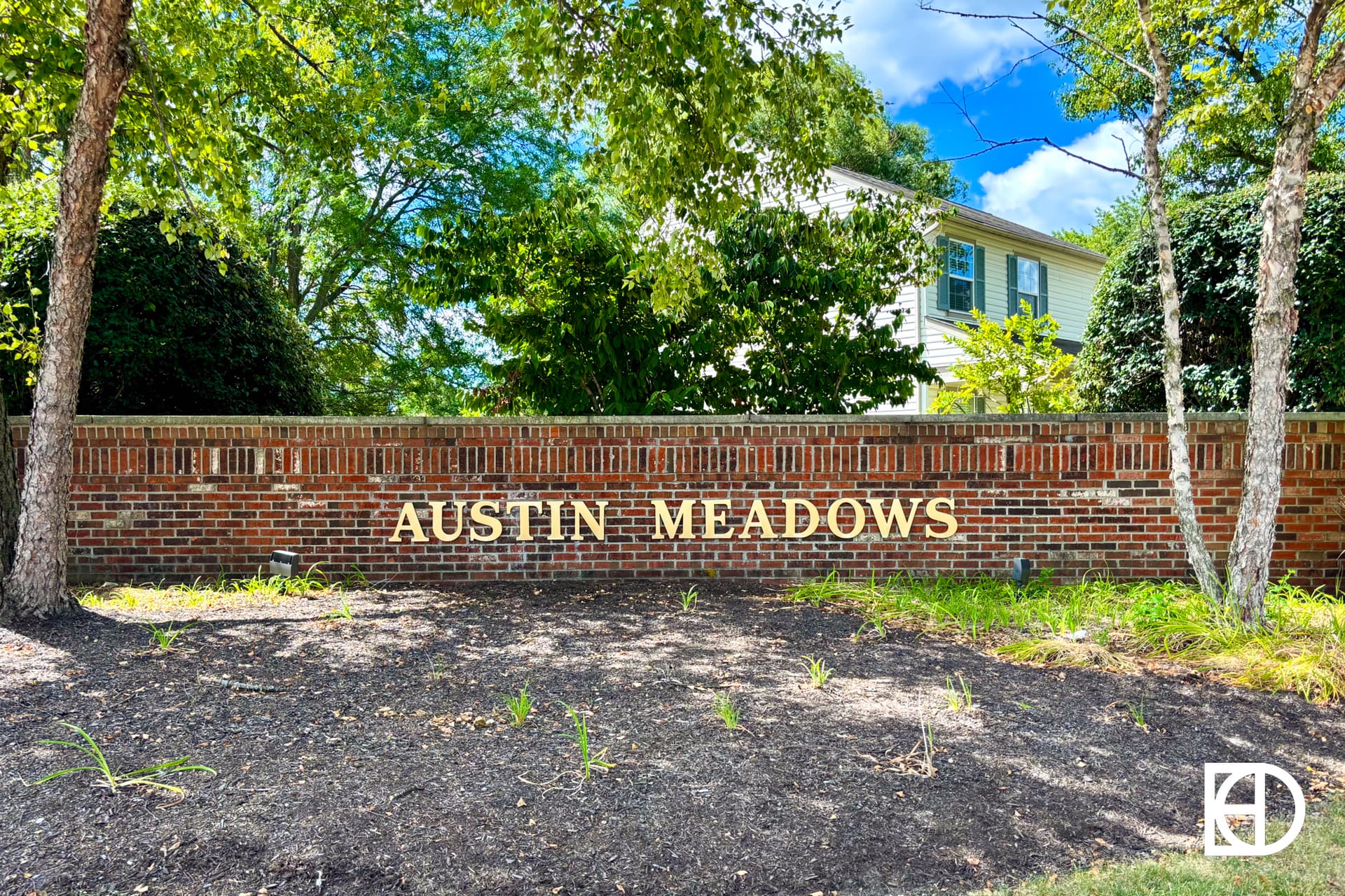 A brick wall sign with gold letters reading AUSTIN MEADOWS in front of trees and a house on a sunny day. A white KD logo is in the bottom right corner.