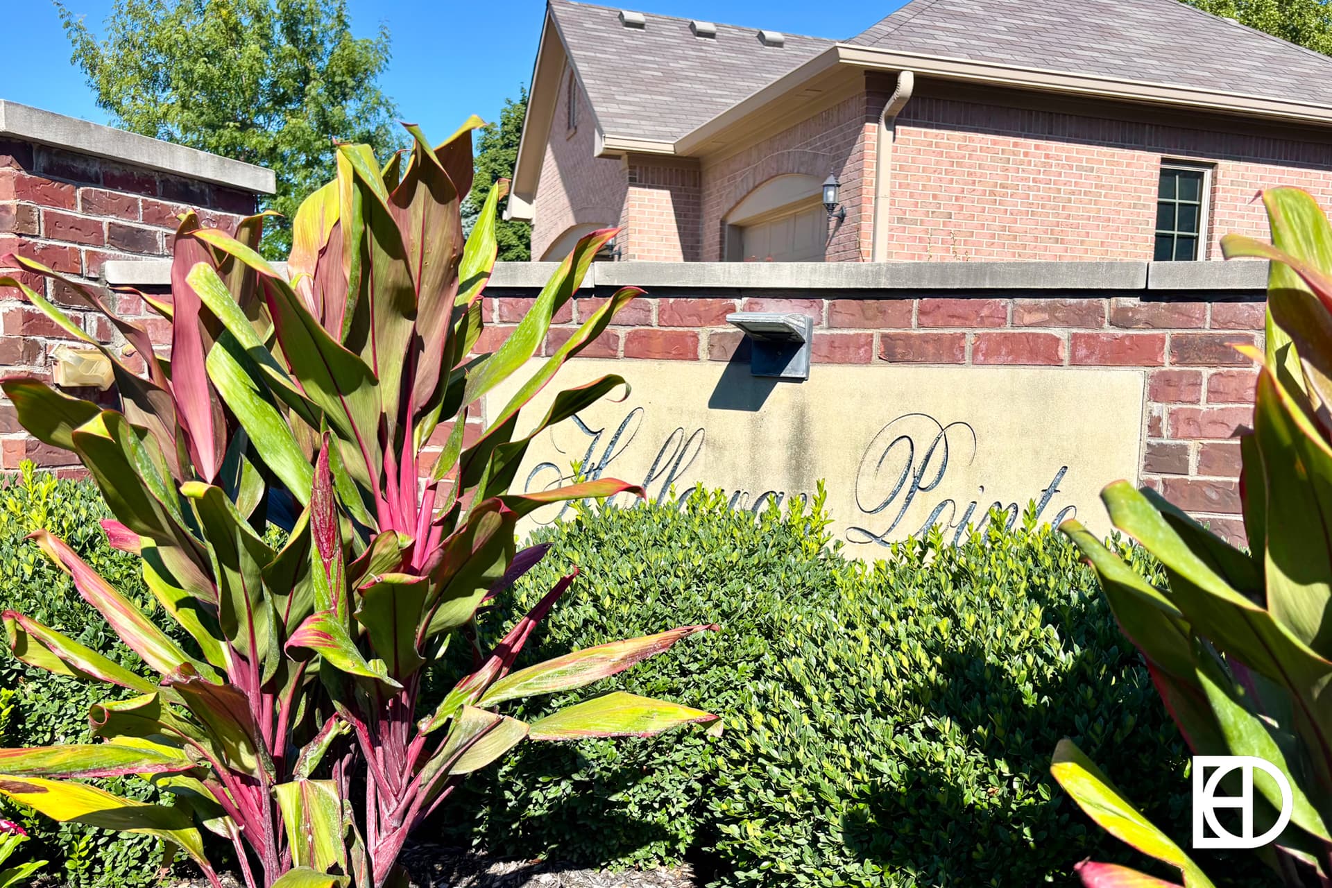 A brick wall with a partially visible sign reading Halloway Point is surrounded by green and red plants; a house with a sloped roof is in the background under a clear blue sky.