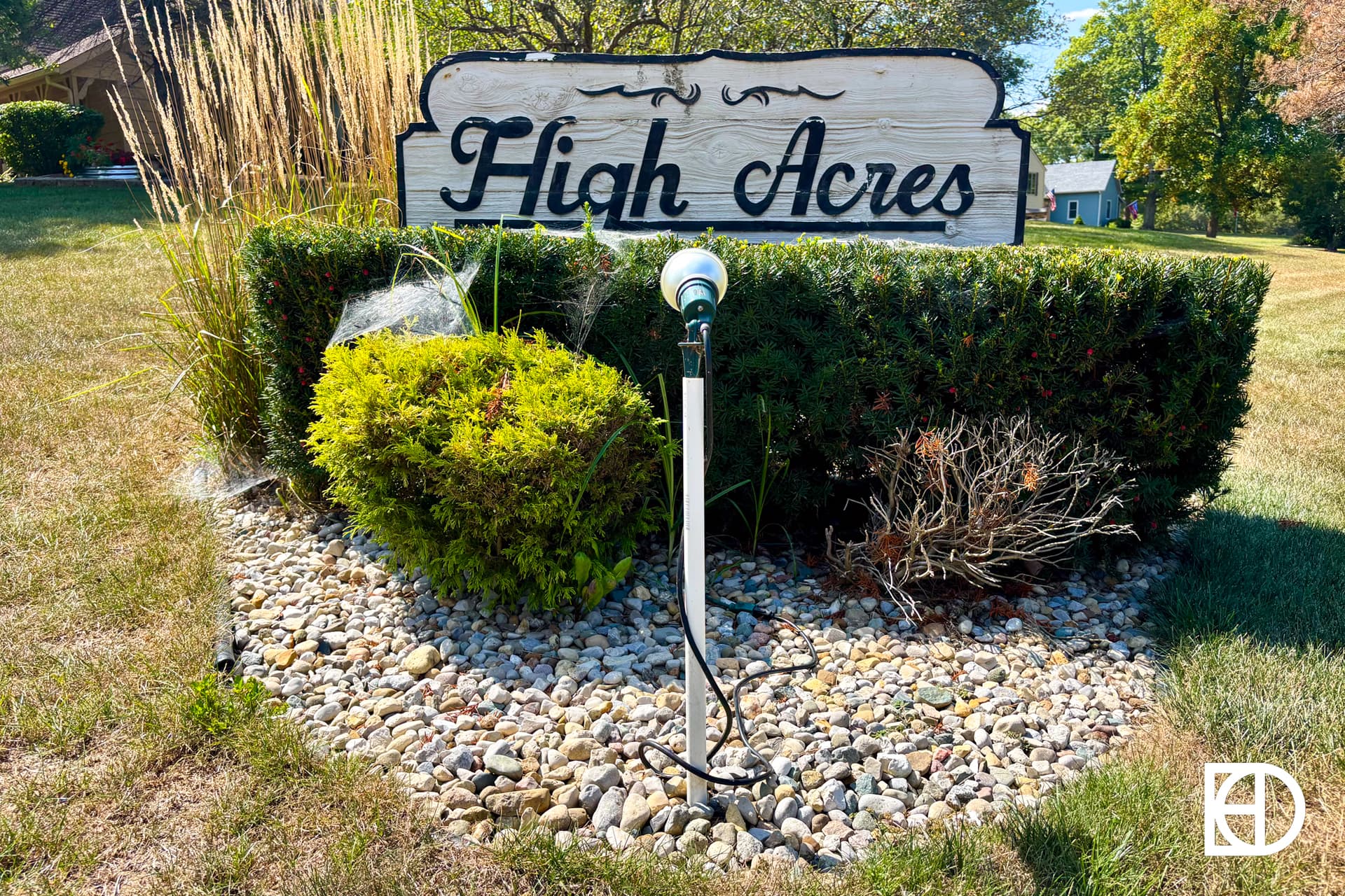 A wooden sign reading High Acres stands in front of a neatly trimmed hedge and rocks, with a garden spotlight and some spider web decorations on the bush. Trees and a house are visible in the background.