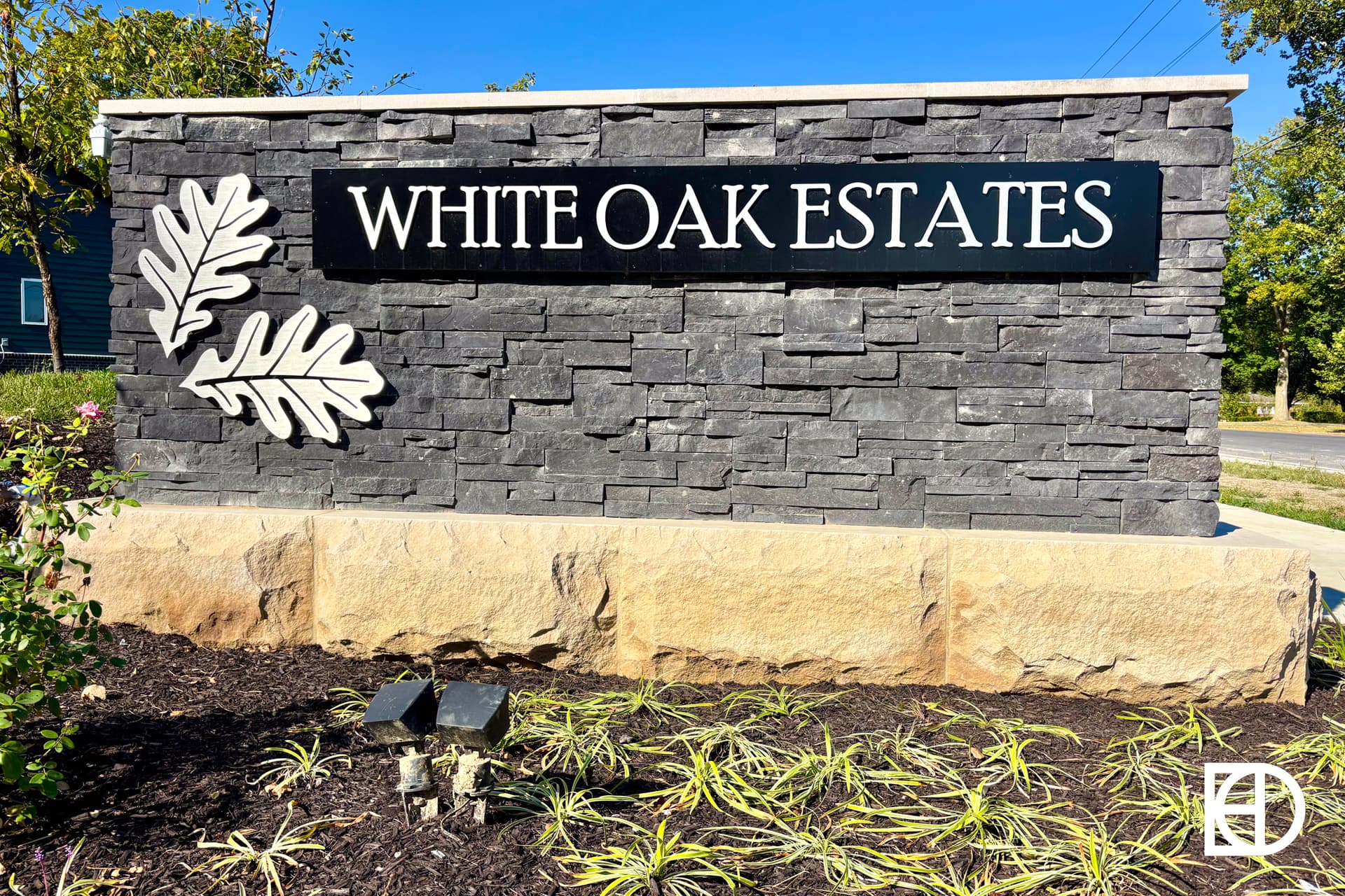 A stone entrance sign reads WHITE OAK ESTATES with two white oak leaf designs on the left, surrounded by landscaping and plants in front.