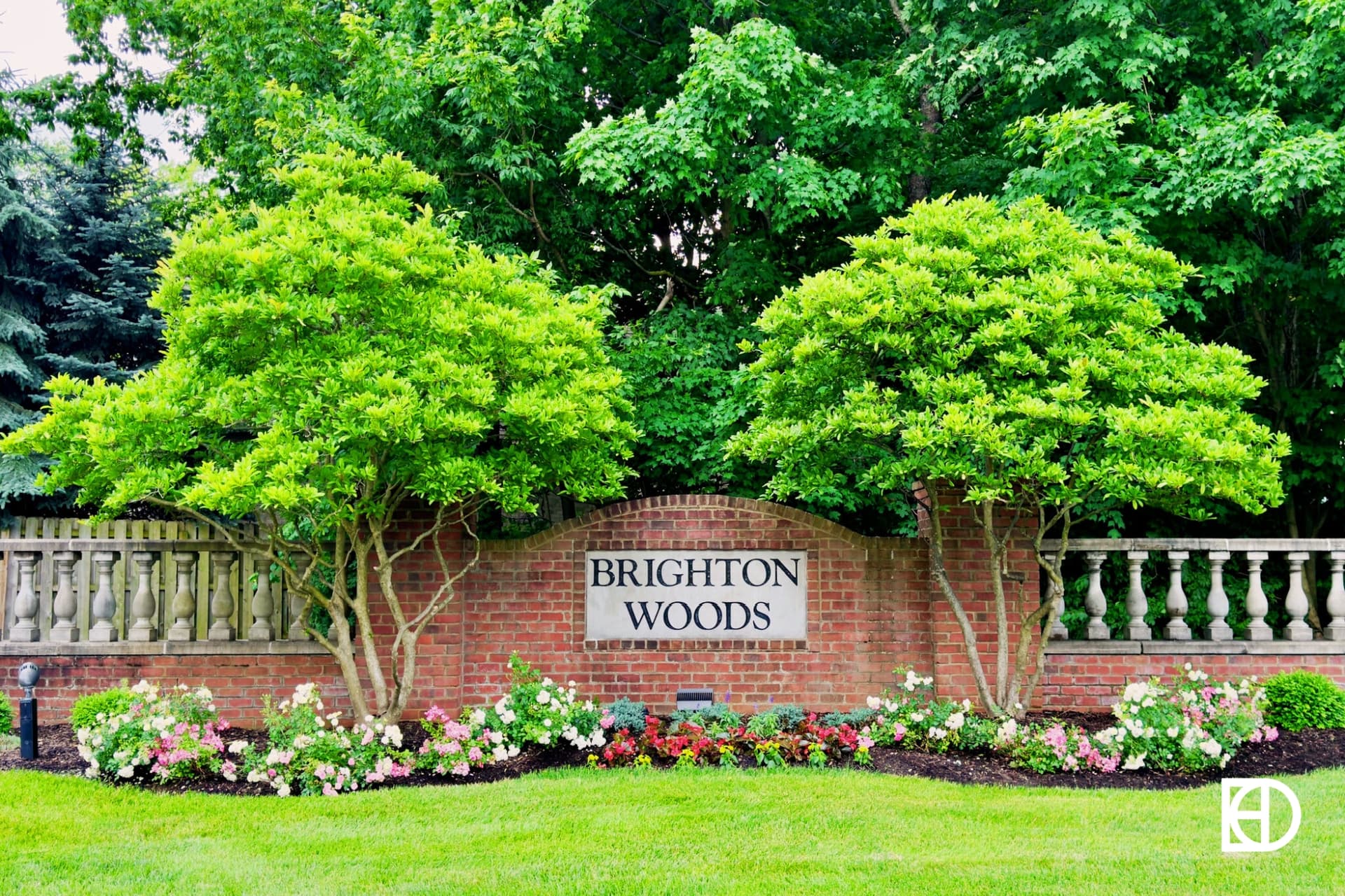 A brick sign reading Brighton Woods is set in front of a landscaped garden with trimmed trees, flowers, and a wooden fence, surrounded by greenery.