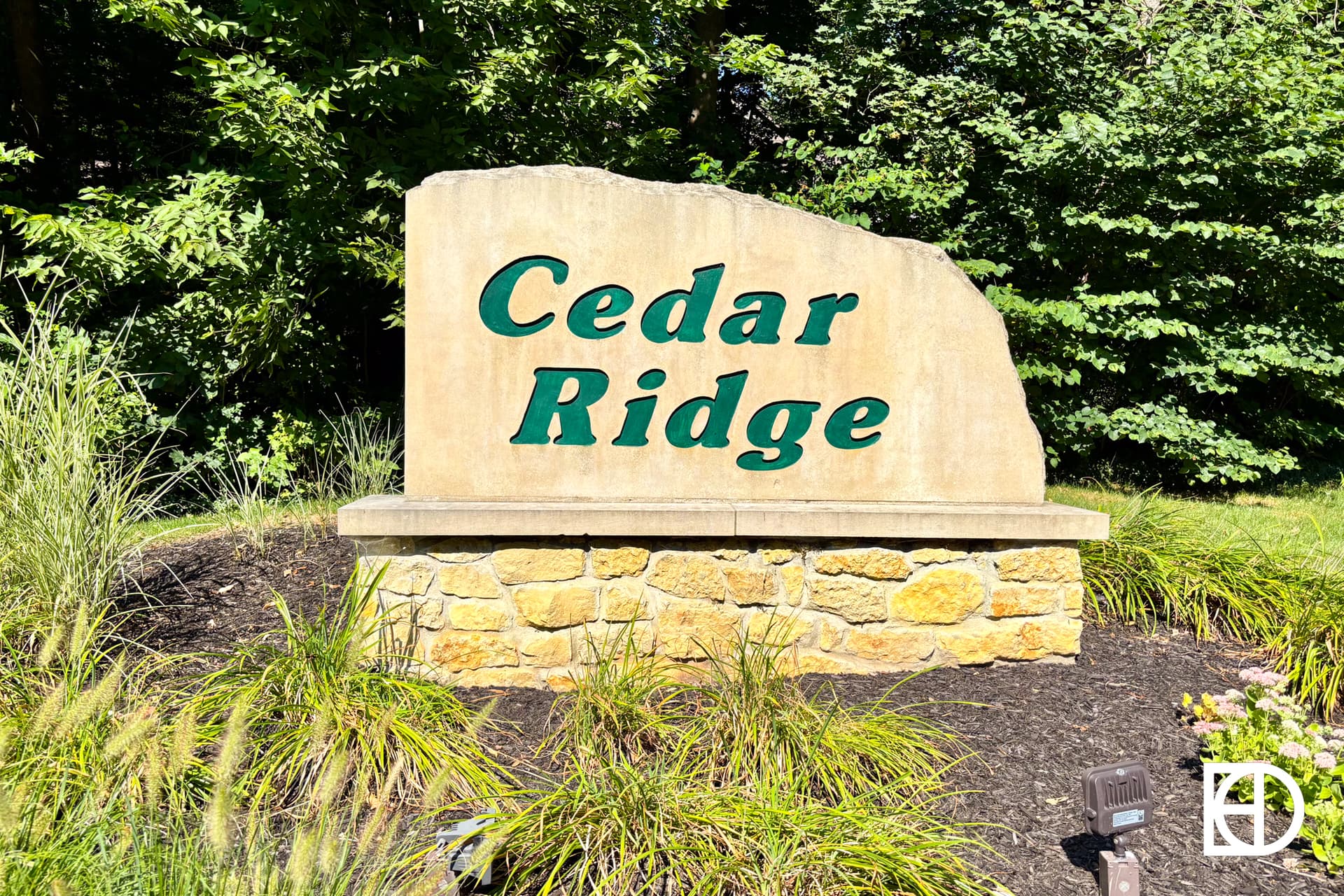 Large stone sign with green letters reading Cedar Ridge, set against a background of trees and greenery, with landscaping and grass in the foreground.