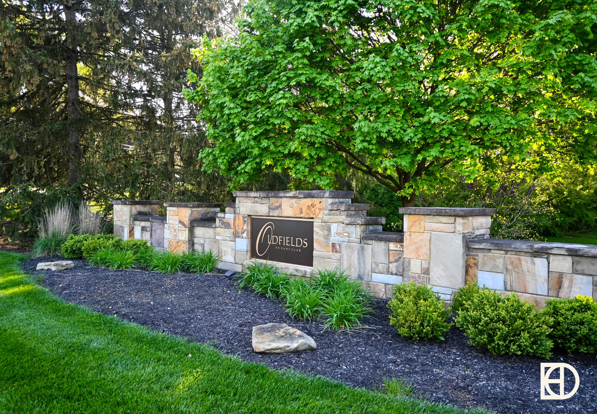 A neighborhood sign displaying "Oldfields on Hunt Club" on a green lawn, with shrubs in front of trees.